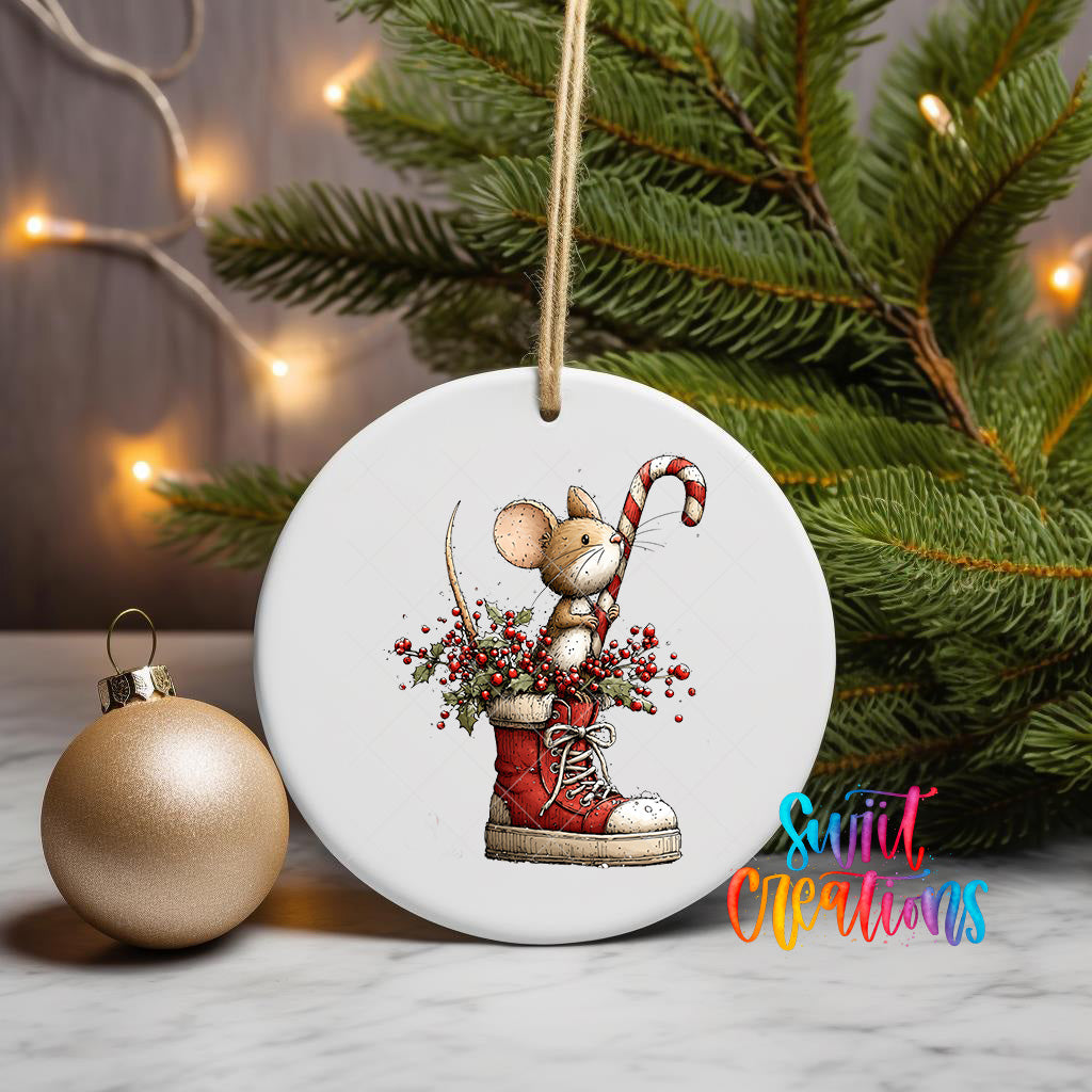 A white ceramic ornament featuring a mouse in a red boot, holding a candy cane, is displayed on a white marble surface next to a gold ornament and a Christmas tree.
