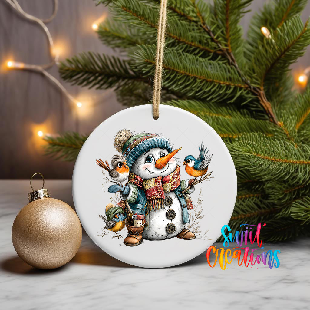 A white ceramic ornament featuring a snowman with a bird perched on its shoulder, surrounded by pine branches and a gold ornament, with a string of lights in the background.