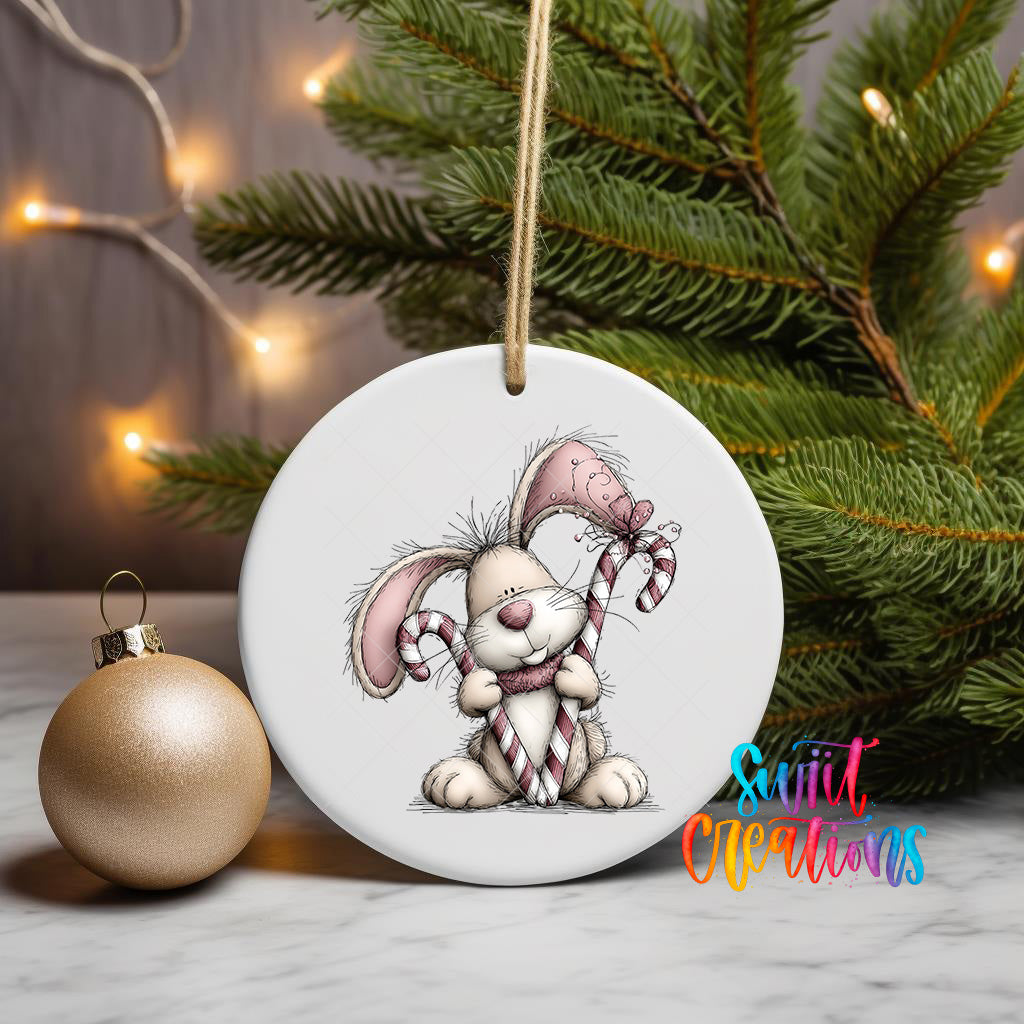 A white ceramic ornament featuring a cartoon rabbit holding a candy cane, placed on a marble surface next to a gold ornament and a Christmas tree with twinkling lights.