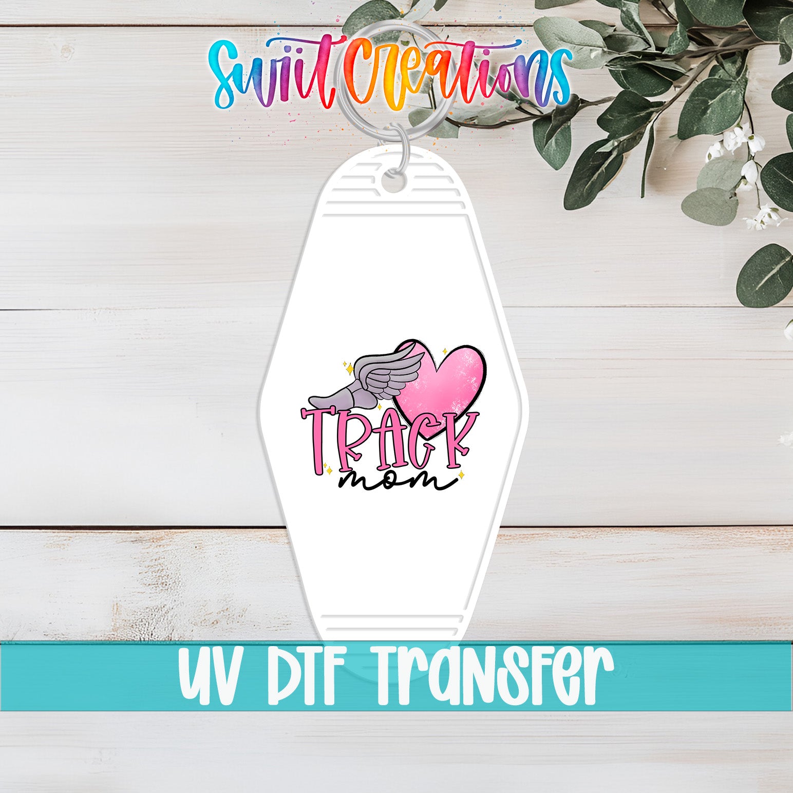 A keychain with a pink heart and the word "TRACK mom" printed on it, along with a colorful logo. The keychain is placed on a wooden surface, and there is a plant in the background.