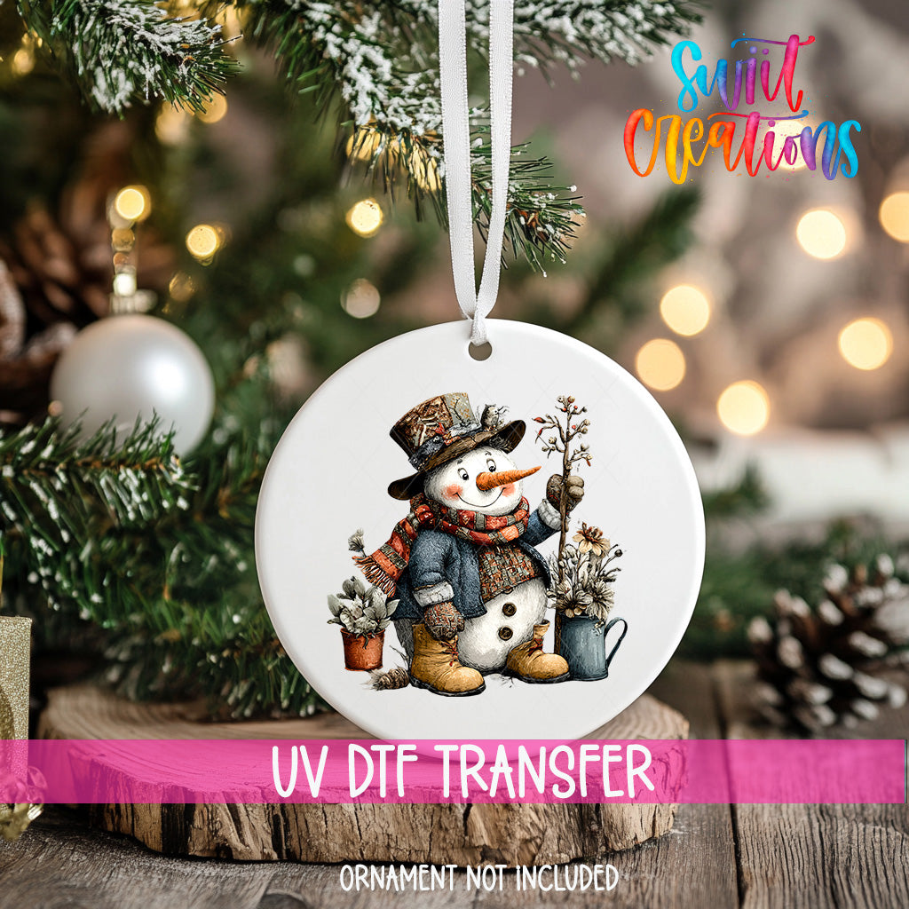 A white ornament with a snowman design hangs on a Christmas tree, surrounded by pine branches and other holiday decorations.