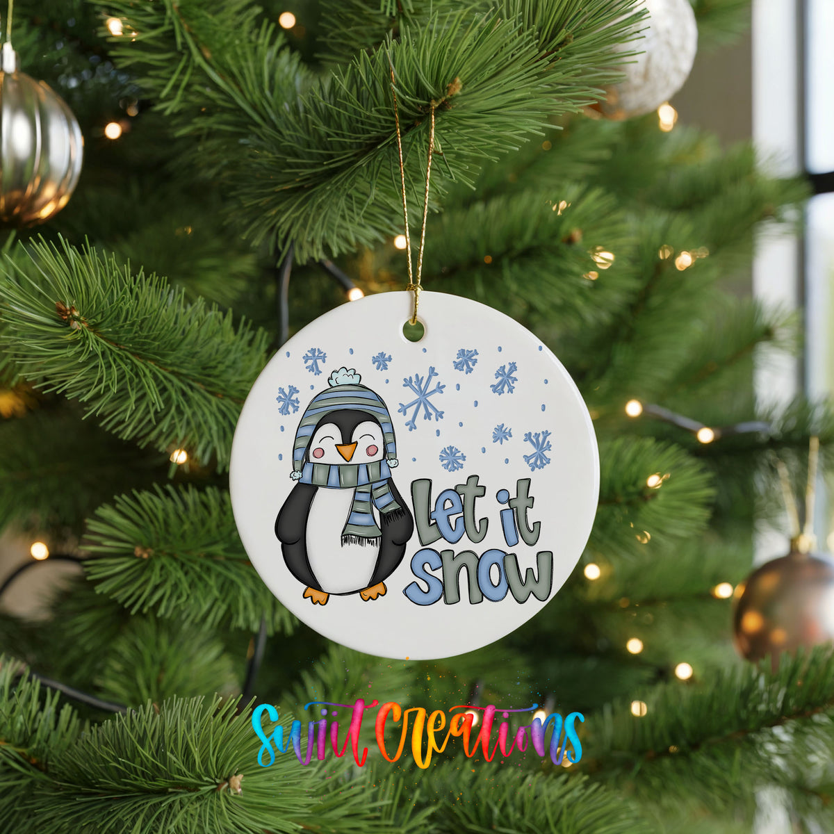 A white ornament with a cartoon penguin wearing a scarf and hat, along with the text "Let it snow" is hanging on a Christmas tree.