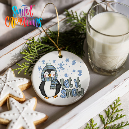 A white ornament with a penguin wearing a scarf and hat, holding a snowflake, and the text "Let it snow" is displayed on a white tray alongside a glass of milk and some cookies.