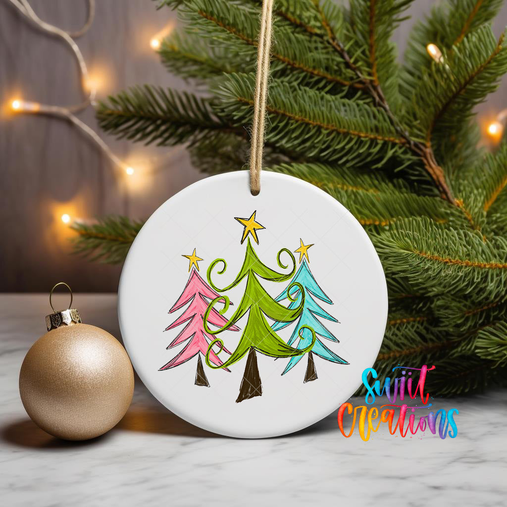 A white ceramic ornament with a colorful illustration of three Christmas trees is displayed on a marble surface, accompanied by a gold ornament and a string of twinkling lights.