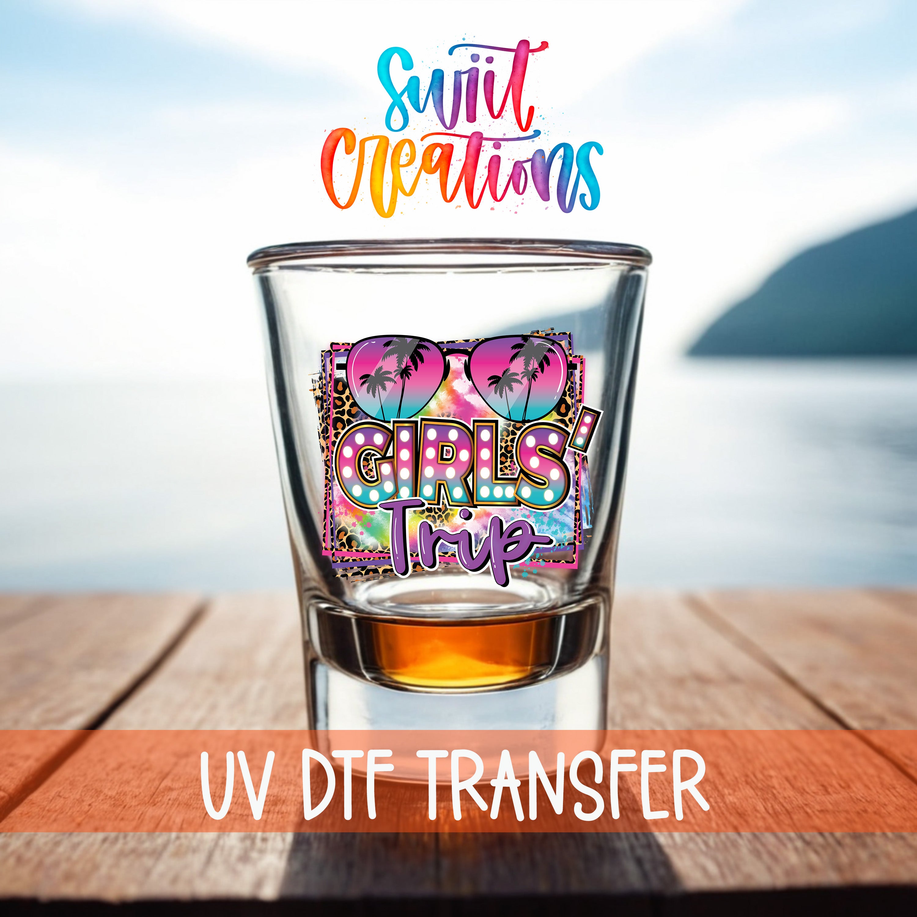 A glass with a colorful graphic design featuring sunglasses and the text "Girls Trip" on it, placed on a wooden surface with a body of water in the background.