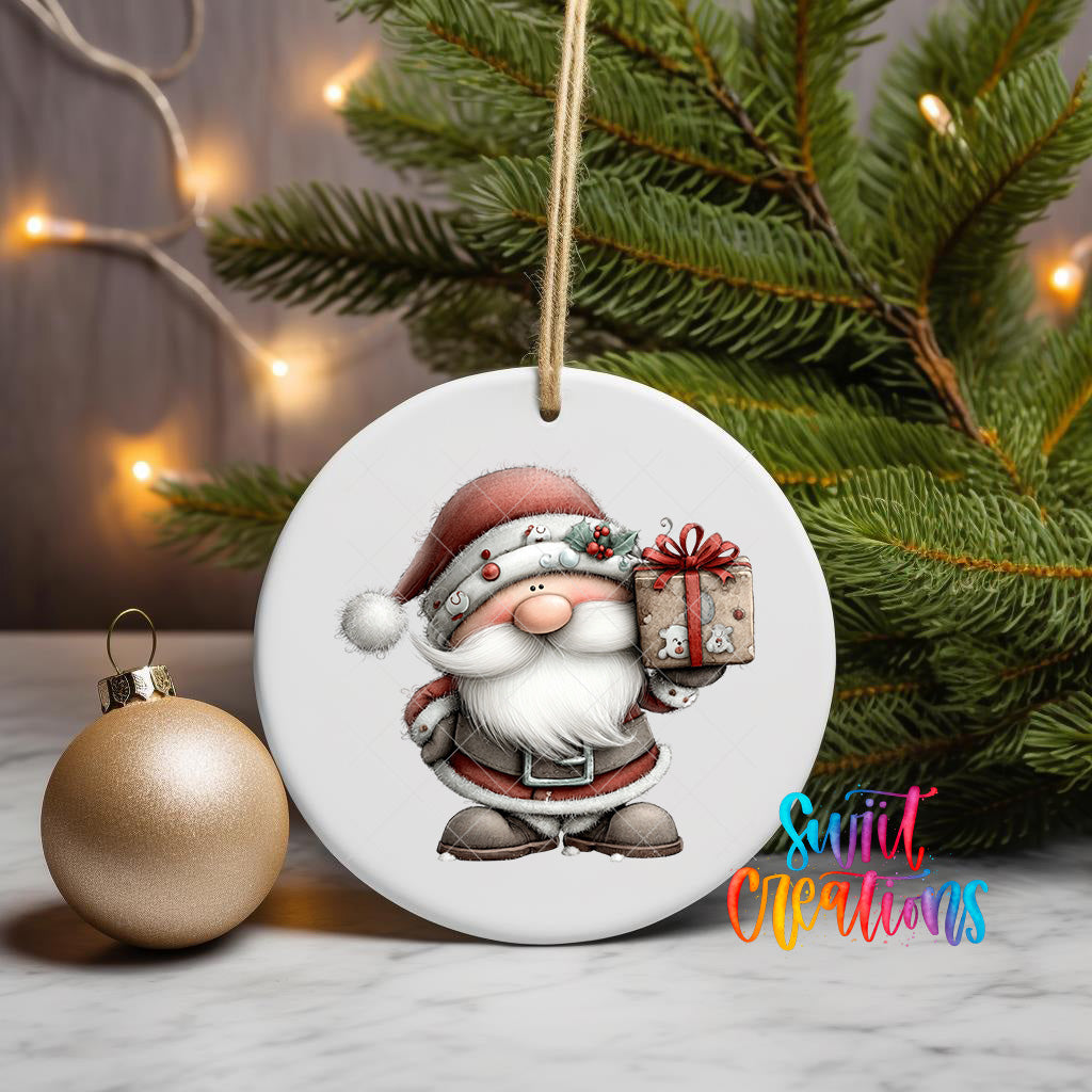 A white ornament featuring a cartoon Santa Claus holding a gift, surrounded by a Christmas tree and a gold ornament, with a blurred background of twinkling lights.