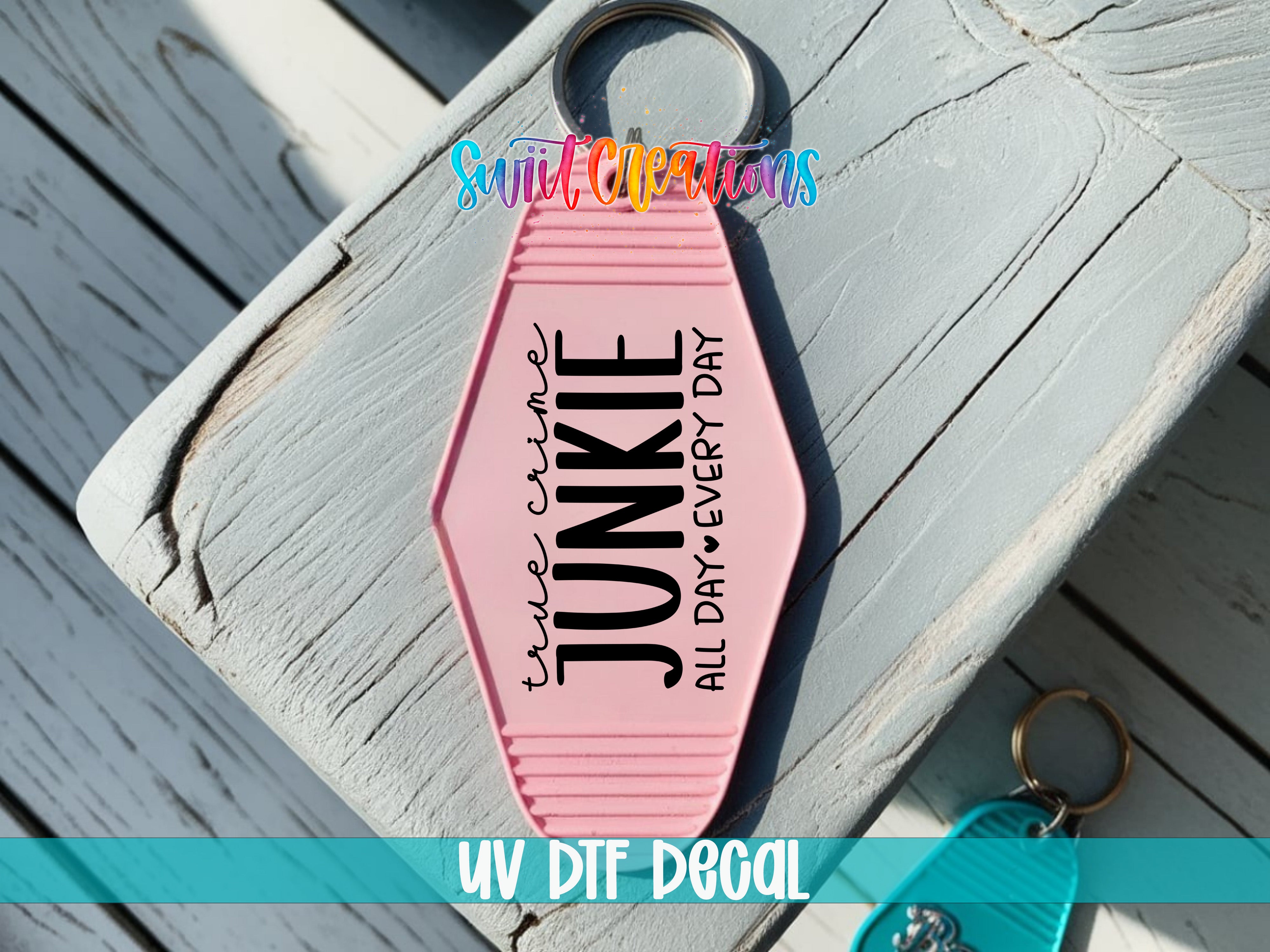 A pink keychain with the text "True Crime Junkie All Day Every Day" printed on it, placed on a wooden surface.