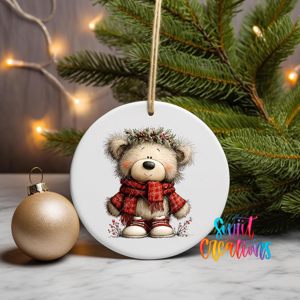 A white ceramic ornament featuring a teddy bear wearing a red scarf and a festive wreath, placed on a marble surface next to a gold ornament and a Christmas tree.