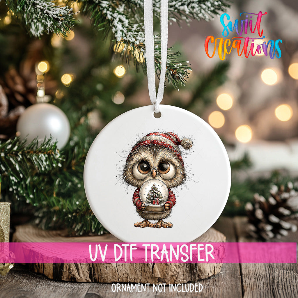 A white ornament with a cute owl wearing a red and white hat, holding a snow globe, is hanging on a Christmas tree.