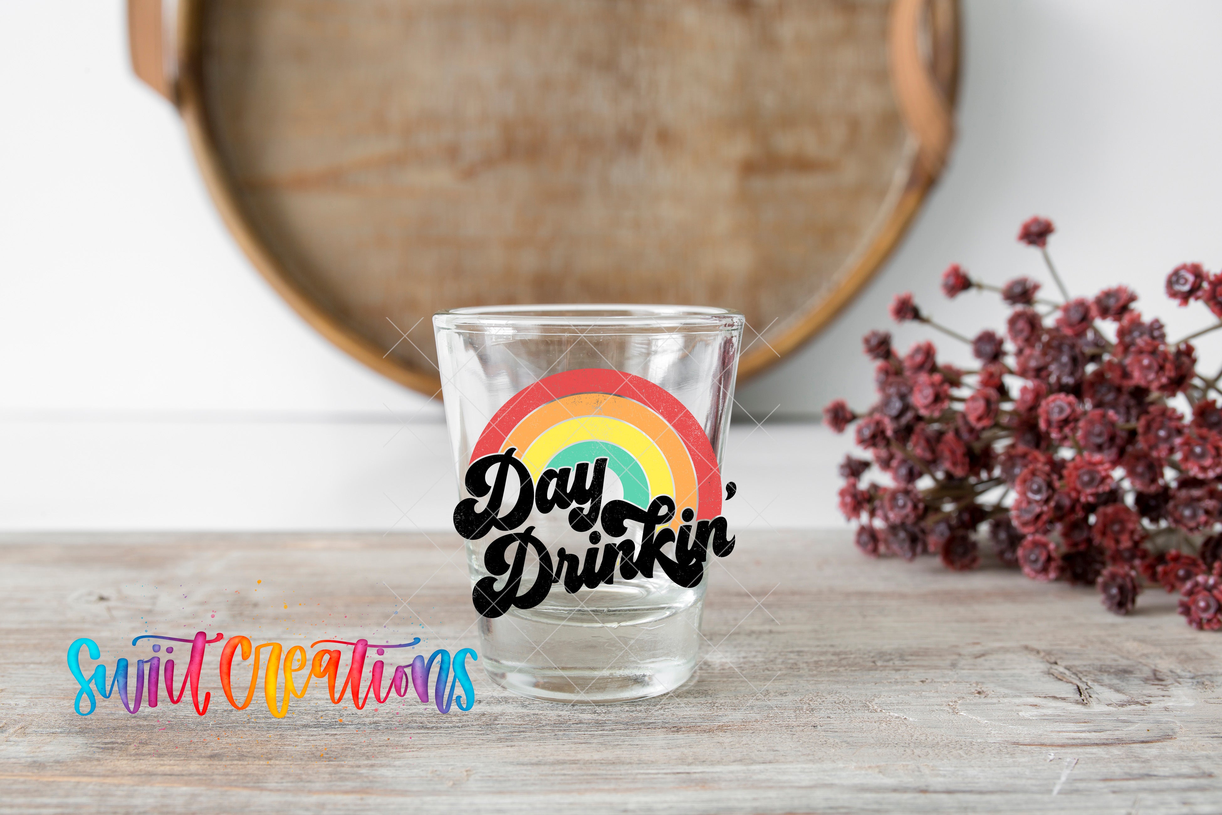 A glass with a rainbow-colored design and the text "Day Drinking" is placed on a wooden surface, with a bunch of red flowers in the background.