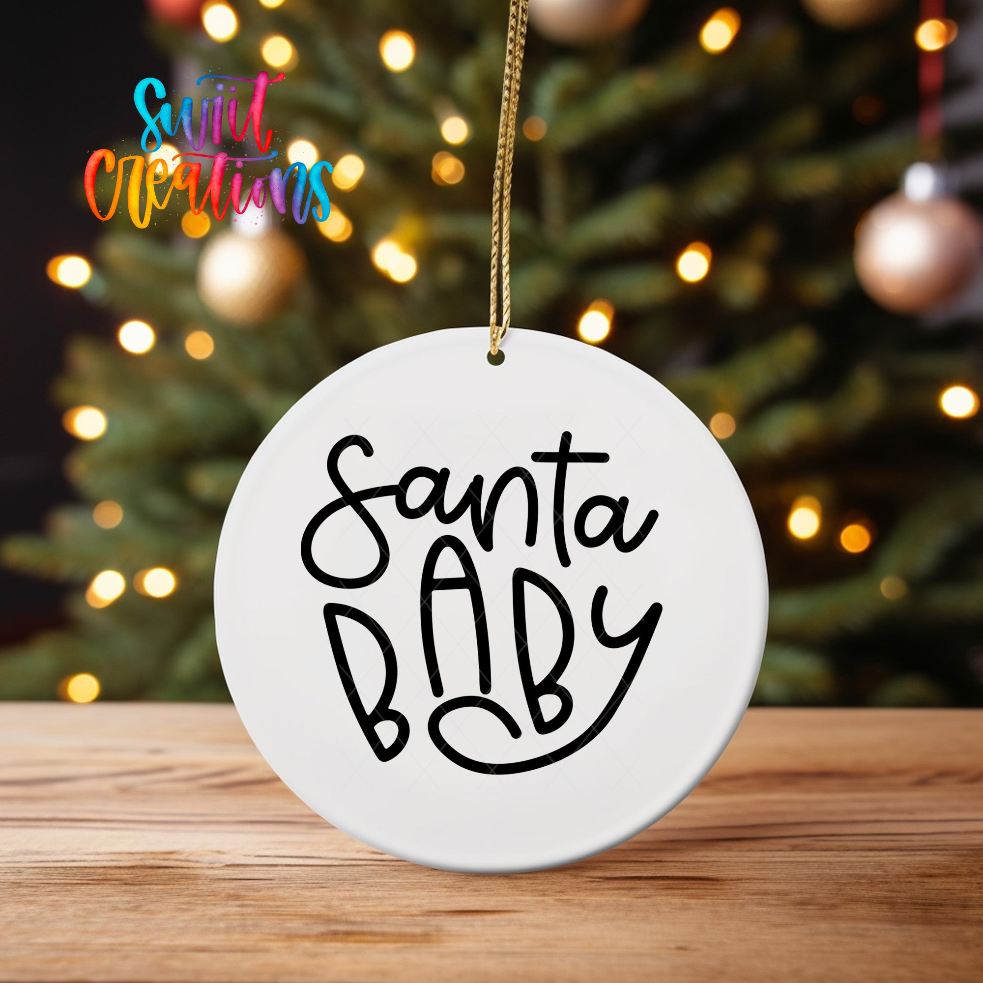 A white ceramic ornament with the text "Santa Baby" written on it, hanging from a string. The ornament is placed on a wooden surface, with a blurred background of a Christmas tree and colorful lights.