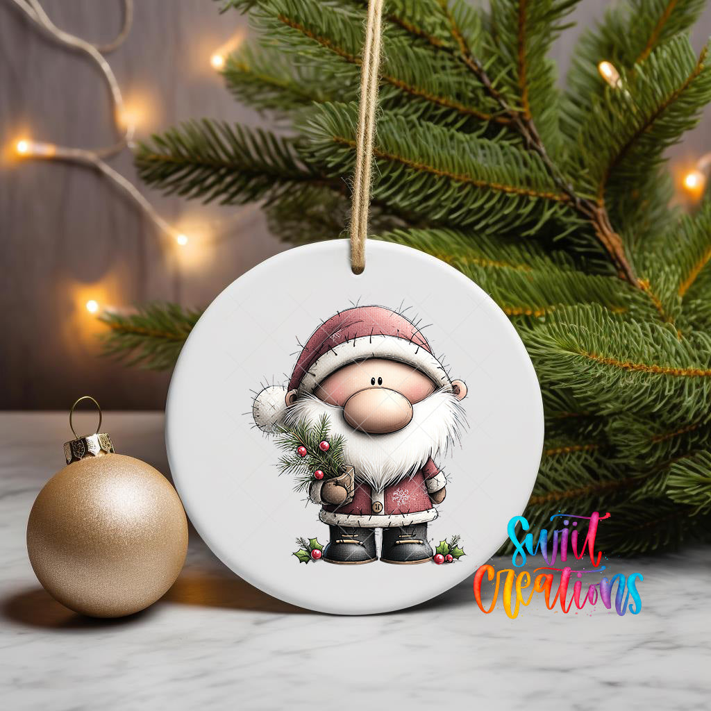 A white ceramic ornament featuring a cartoon Santa Claus holding a pine branch and berries, with a gold ornament to the left and a Christmas tree in the background.