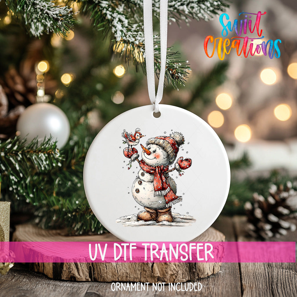 A white ornament with a snowman and a bird is hanging on a Christmas tree, with a pink ribbon around it.