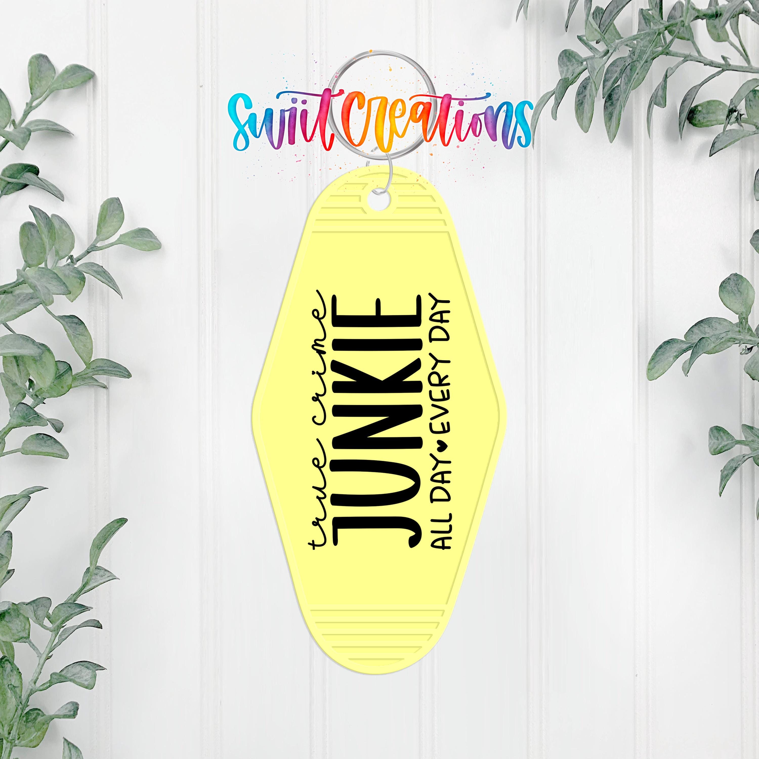A yellow keychain with the text "True Crime Junkie All Day Every Day" printed on it, hanging on a white wall with greenery in the background.