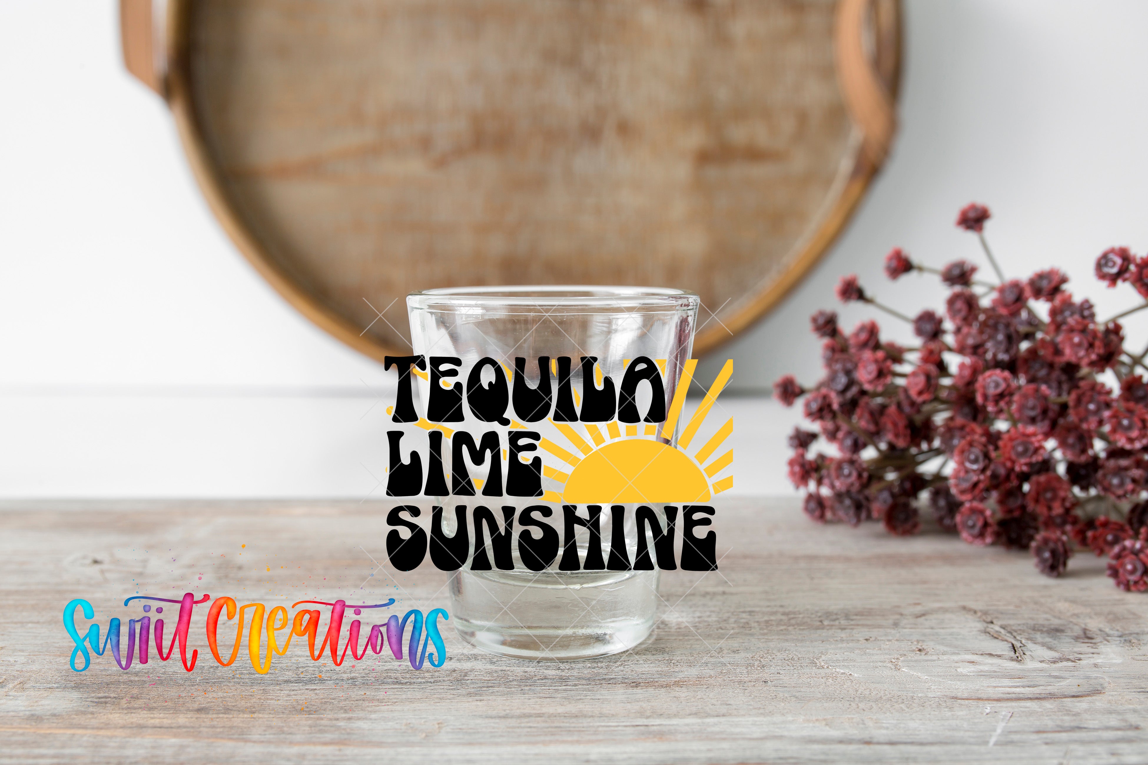 A glass with the text "Tequila Lime Sunshine" printed on it, placed on a wooden surface next to a bunch of red flowers.