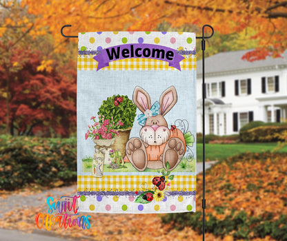 A colorful garden sign featuring a cartoon rabbit and flowers, with the word "Welcome" displayed on it. The sign is hanging on a metal pole, and there is a house and trees in the background.