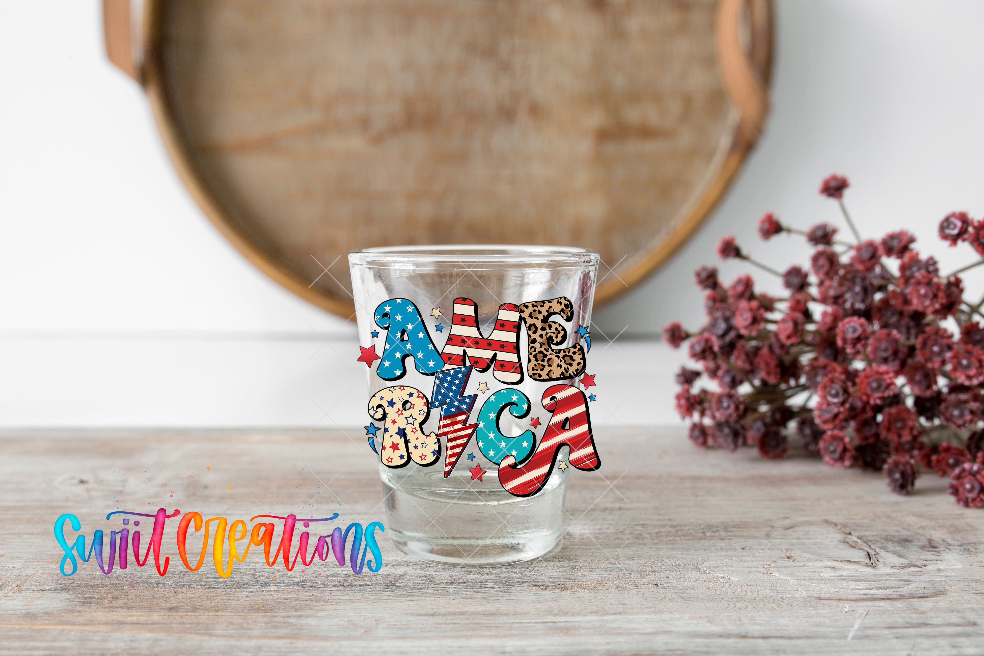 A glass with the word "AMERICA" printed on it, placed on a wooden surface next to a bunch of red flowers.