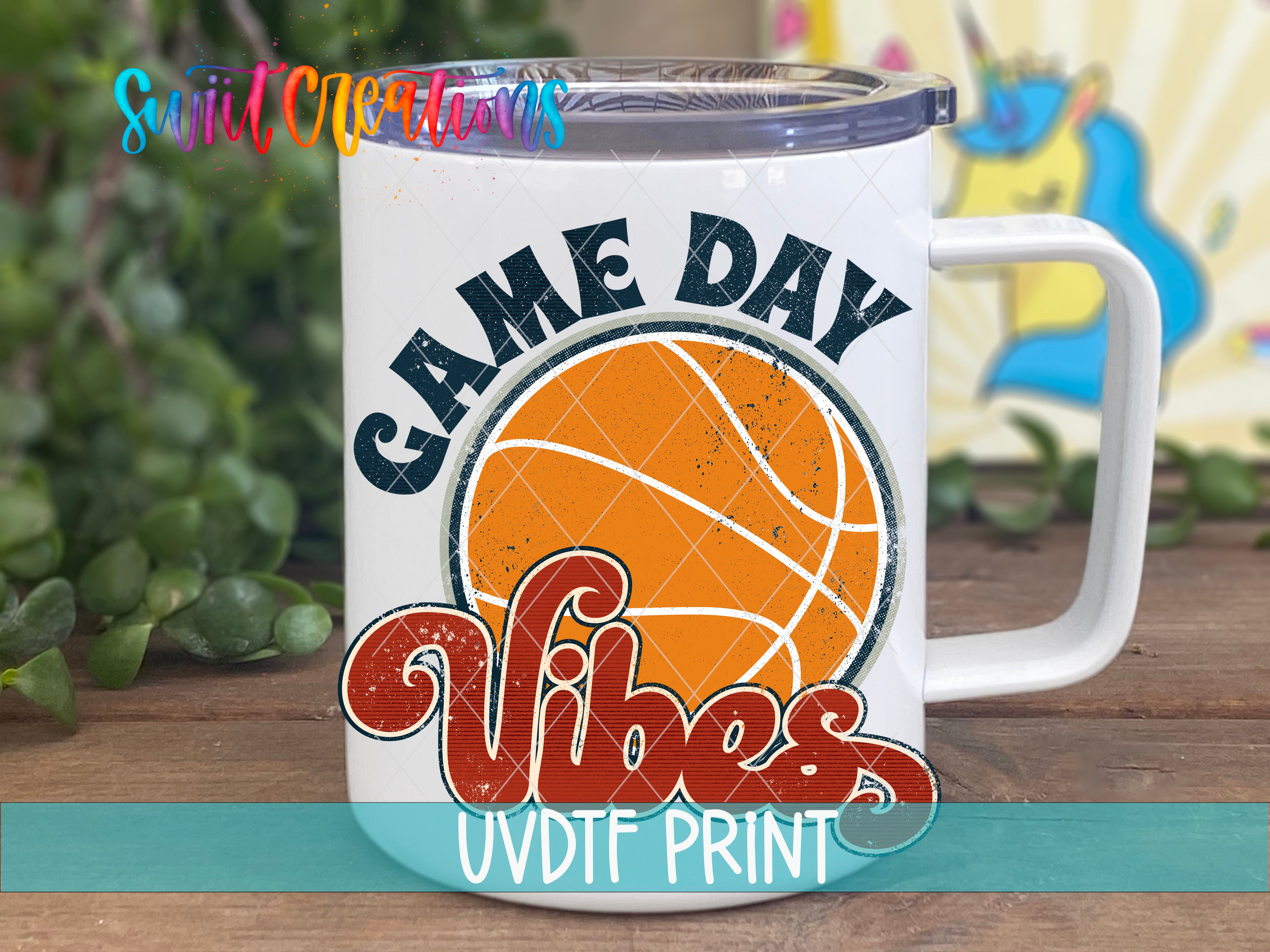 A white mug with a basketball graphic and the text "GAME DAY VIBES" printed on it, placed on a wooden surface with greenery in the background.