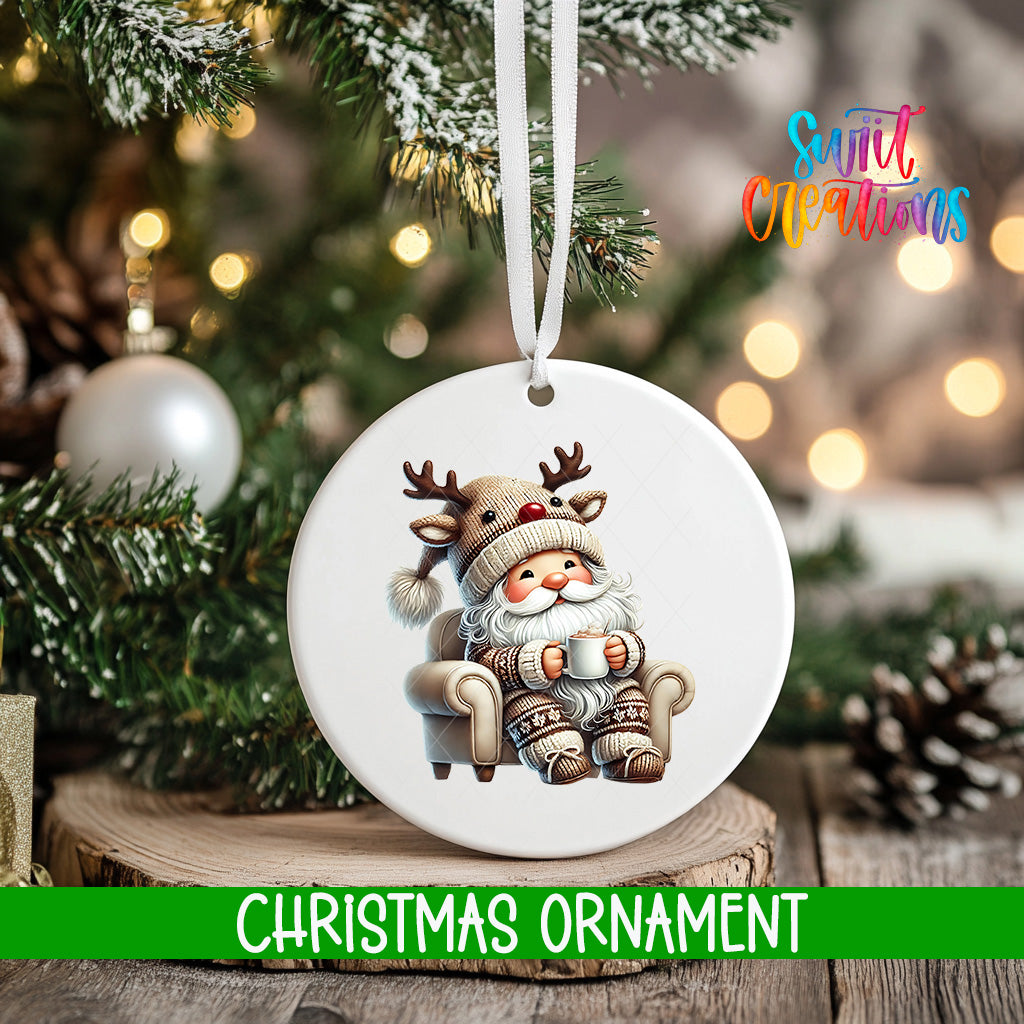 A white Christmas ornament featuring a reindeer sitting in a chair, holding a mug, is hanging on a tree branch.
