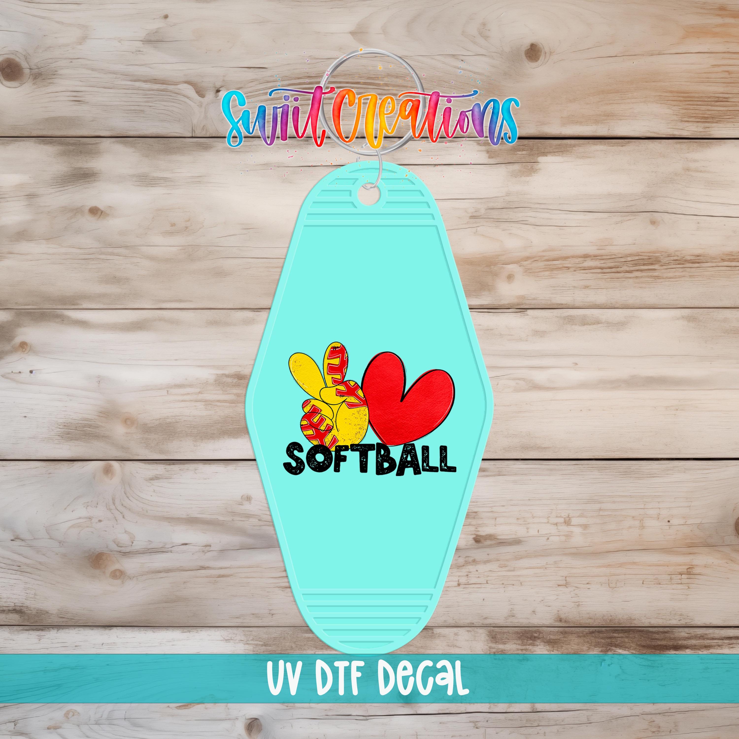 A turquoise keychain with a peace sign and a heart symbol, along with the text "SOFTBALL" and "UV Dtf Decal" printed on it, is displayed on a wooden surface.