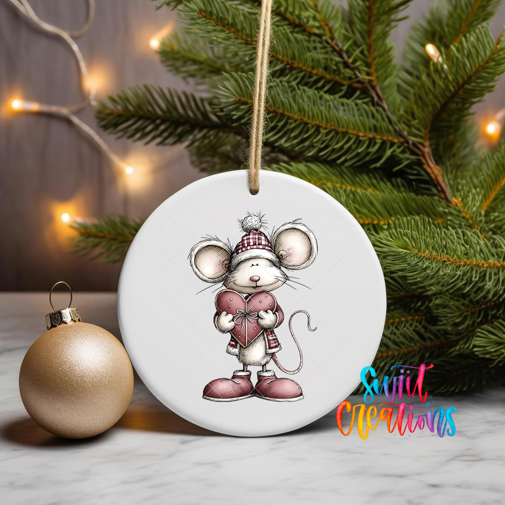 A white ceramic ornament featuring a cartoon mouse wearing a red coat and hat, holding a heart, is displayed on a white surface next to a gold ornament and a Christmas tree with twinkling lights.