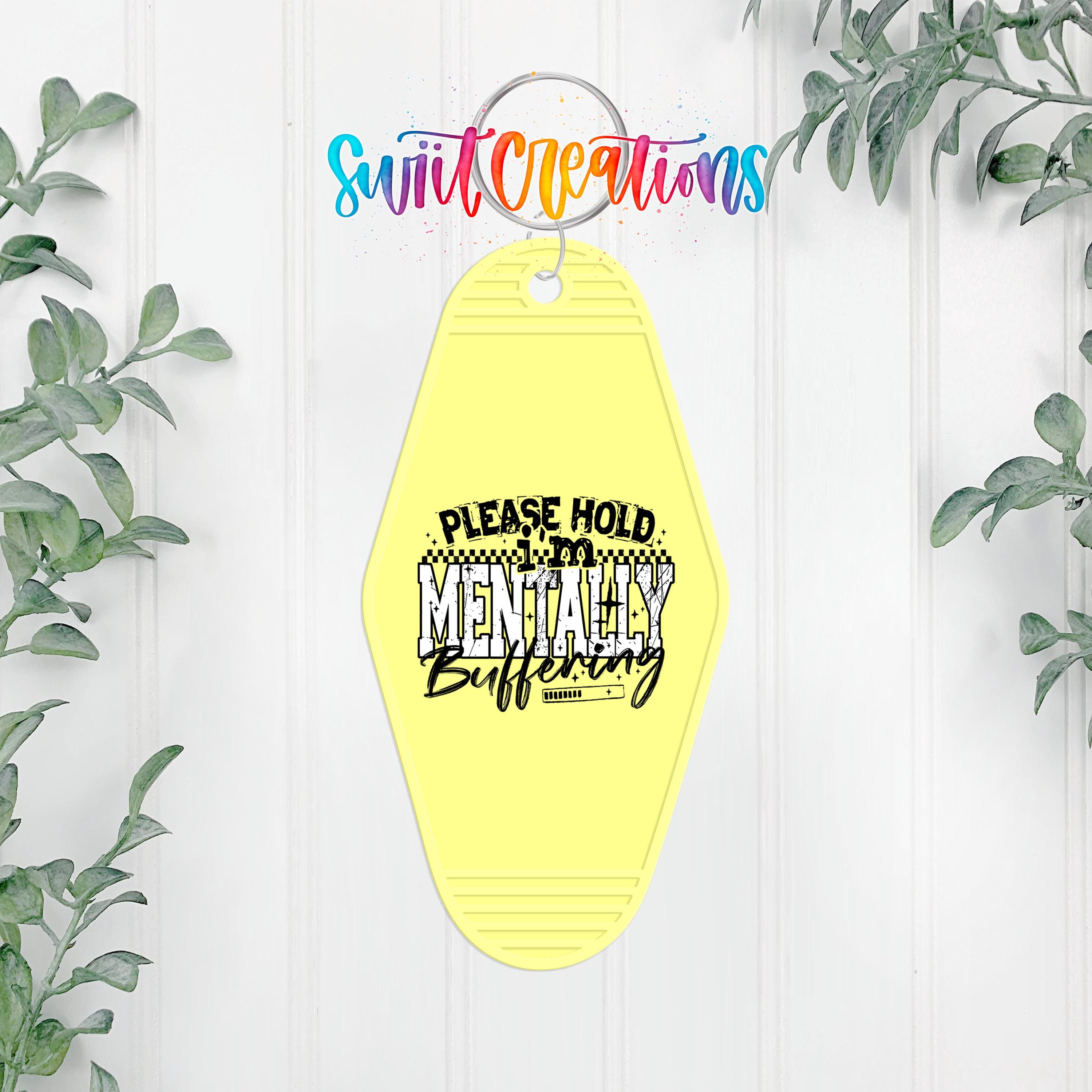 A yellow keychain with a humorous text message that reads "PLEASE HOLD MENTALLY Buffing" is hanging on a white wall, surrounded by greenery.