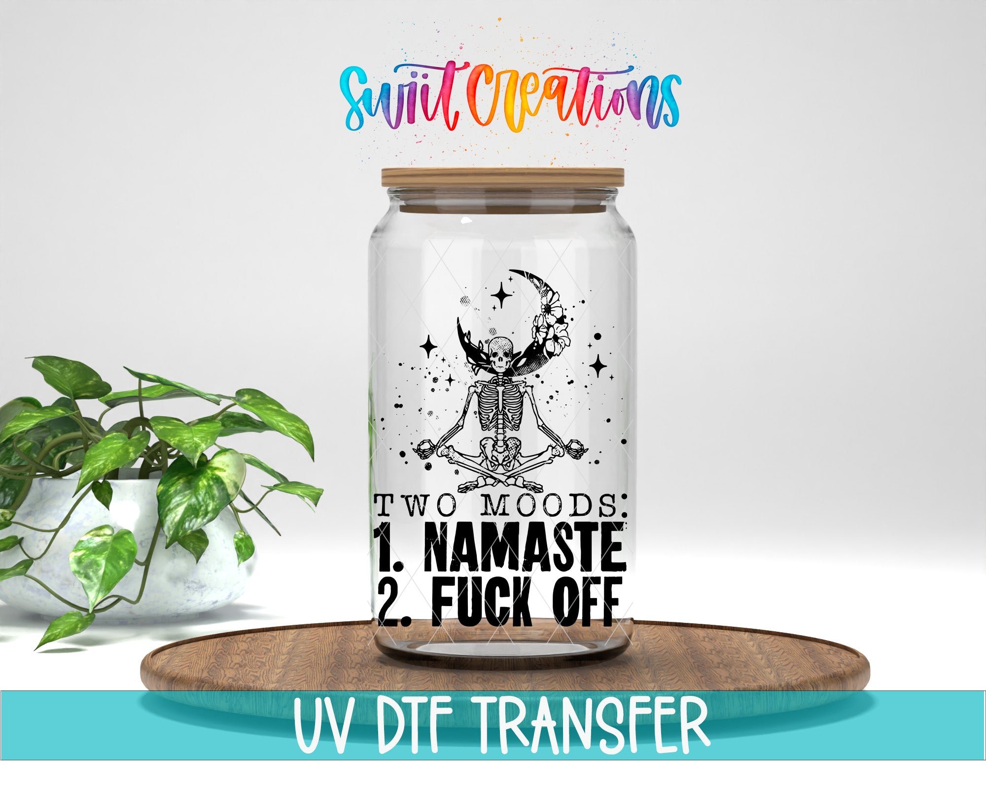 A glass jar with a skeleton and a crescent moon design, along with the text "TWO MOODS: 1. NAMASTE 2. FUCK OFF" printed on it. The jar is placed on a wooden surface, and there is a potted plant in the background.