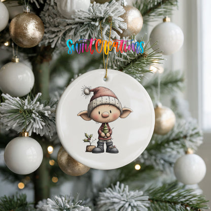 A white ceramic ornament featuring a cartoon elf wearing a Santa hat and holding a Christmas tree, hanging on a decorated Christmas tree.