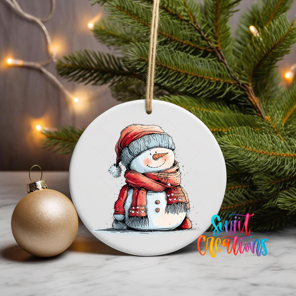 A white ceramic ornament featuring a snowman is displayed on a table, with a gold ornament and a Christmas tree in the background.