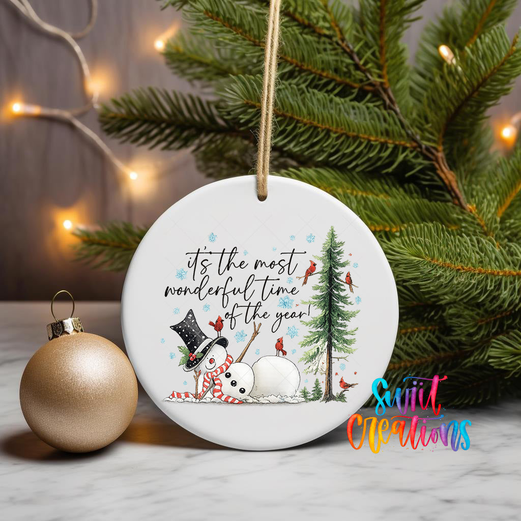 A white ornament with a snowman and Christmas tree design, along with the text "It's the most wonderful time of the year!" and a small gold ornament, is displayed on a table with a Christmas tree in the background.