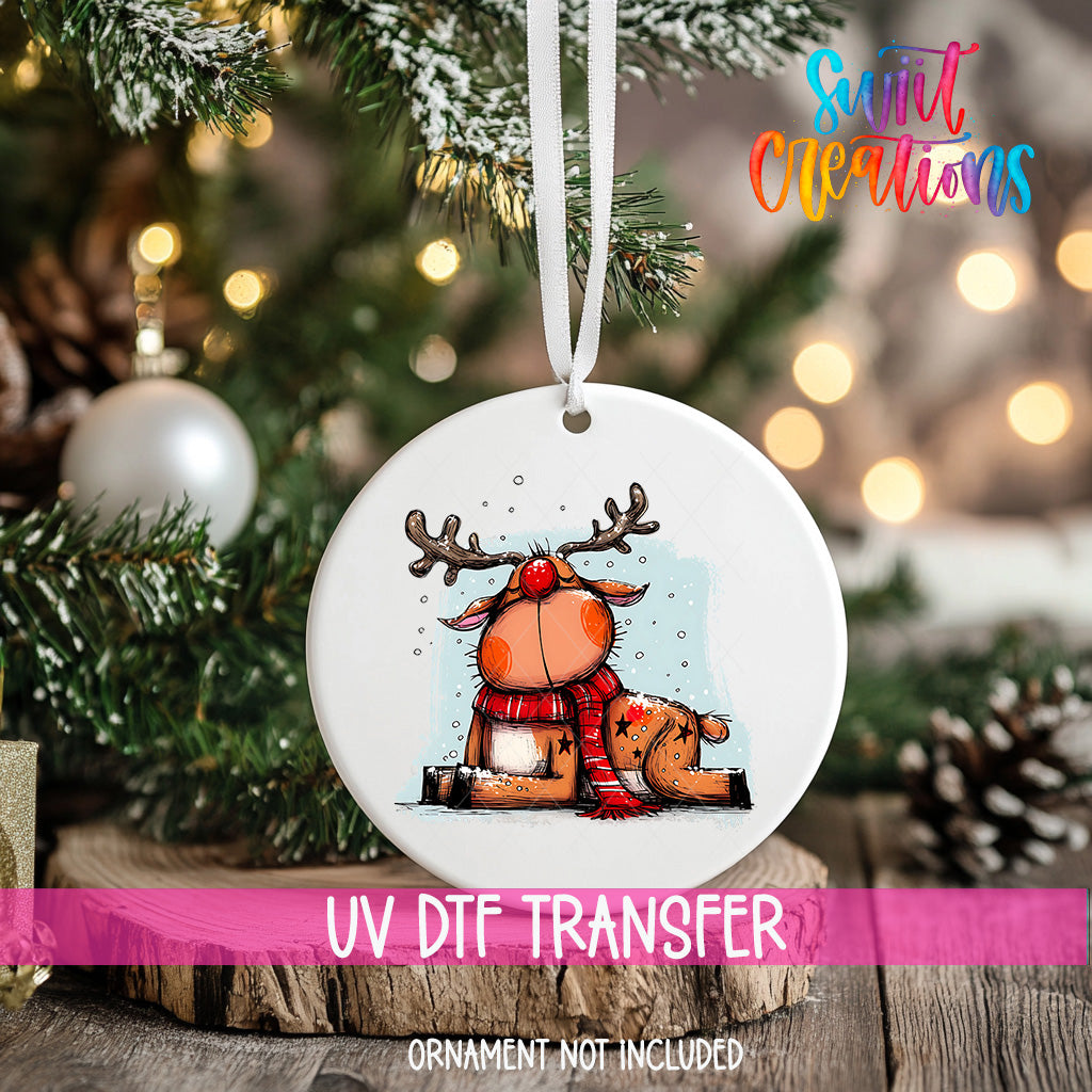 A white ornament with a cartoon reindeer and a red scarf is hanging on a Christmas tree.