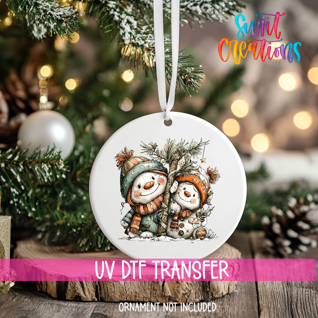 A white ornament with a snowman and pine tree design hangs on a Christmas tree, with a pink ribbon around it.