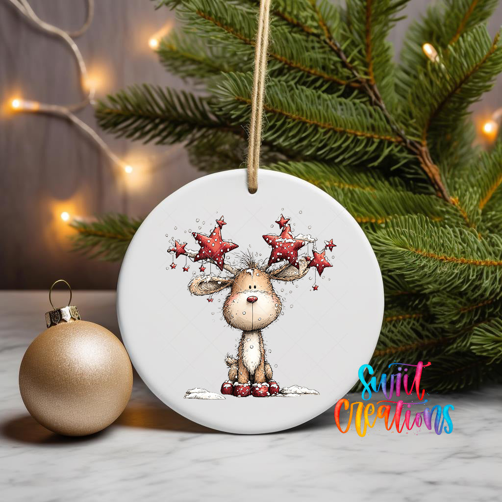 A white ornament with a cartoon reindeer and stars is displayed on a table, accompanied by a gold ornament and a Christmas tree with twinkling lights in the background.