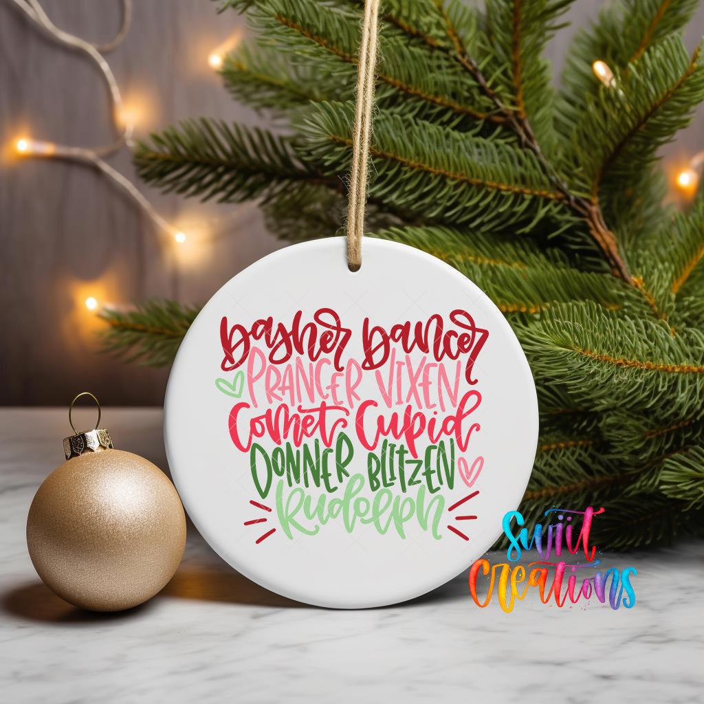 A white ceramic ornament with colorful text that reads "BAYER DANCER PRINCER VIXEN CONNET CUPID DONNER BLIZZEN RUDELEN" is displayed on a white marble surface, with a gold ornament and a Christmas tree in the background.