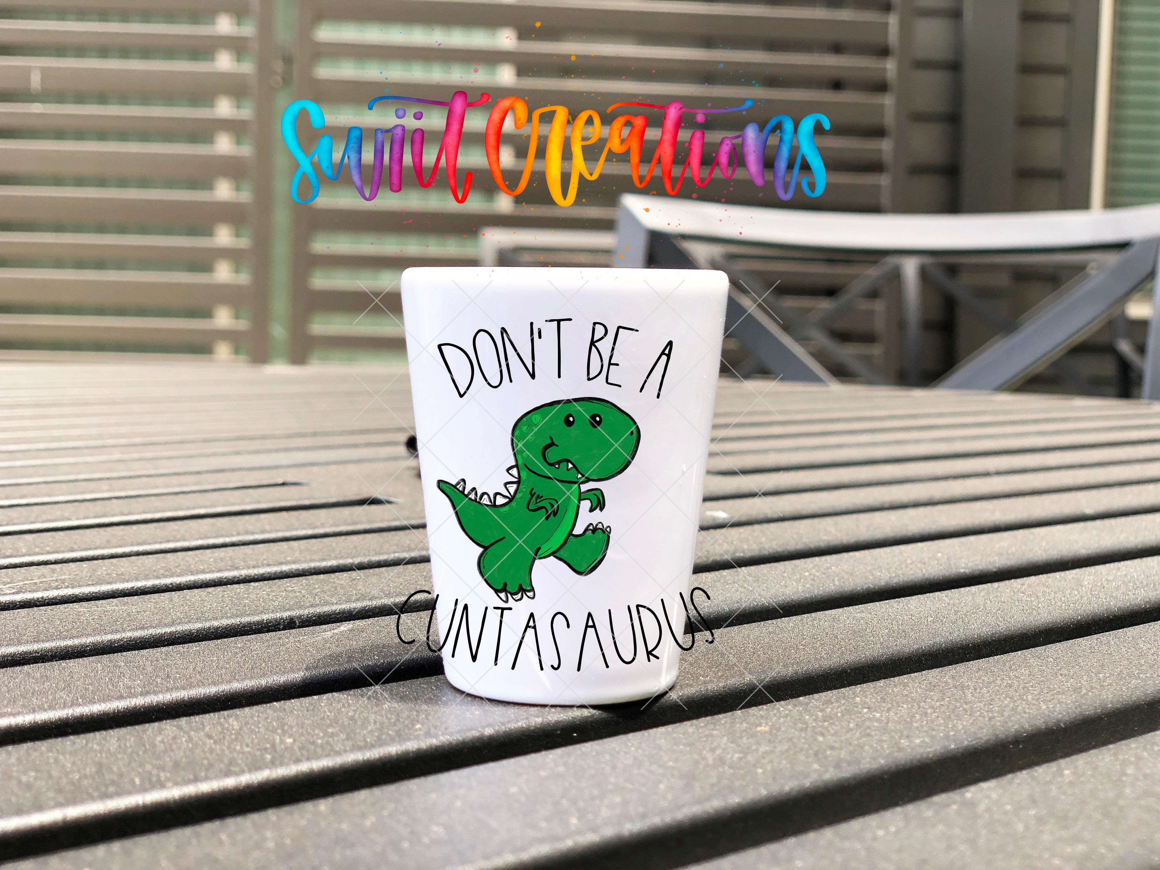 A white cup with a cartoon dinosaur and the text "DON'T BE A CUTASAURUS" printed on it, placed on a wooden surface.