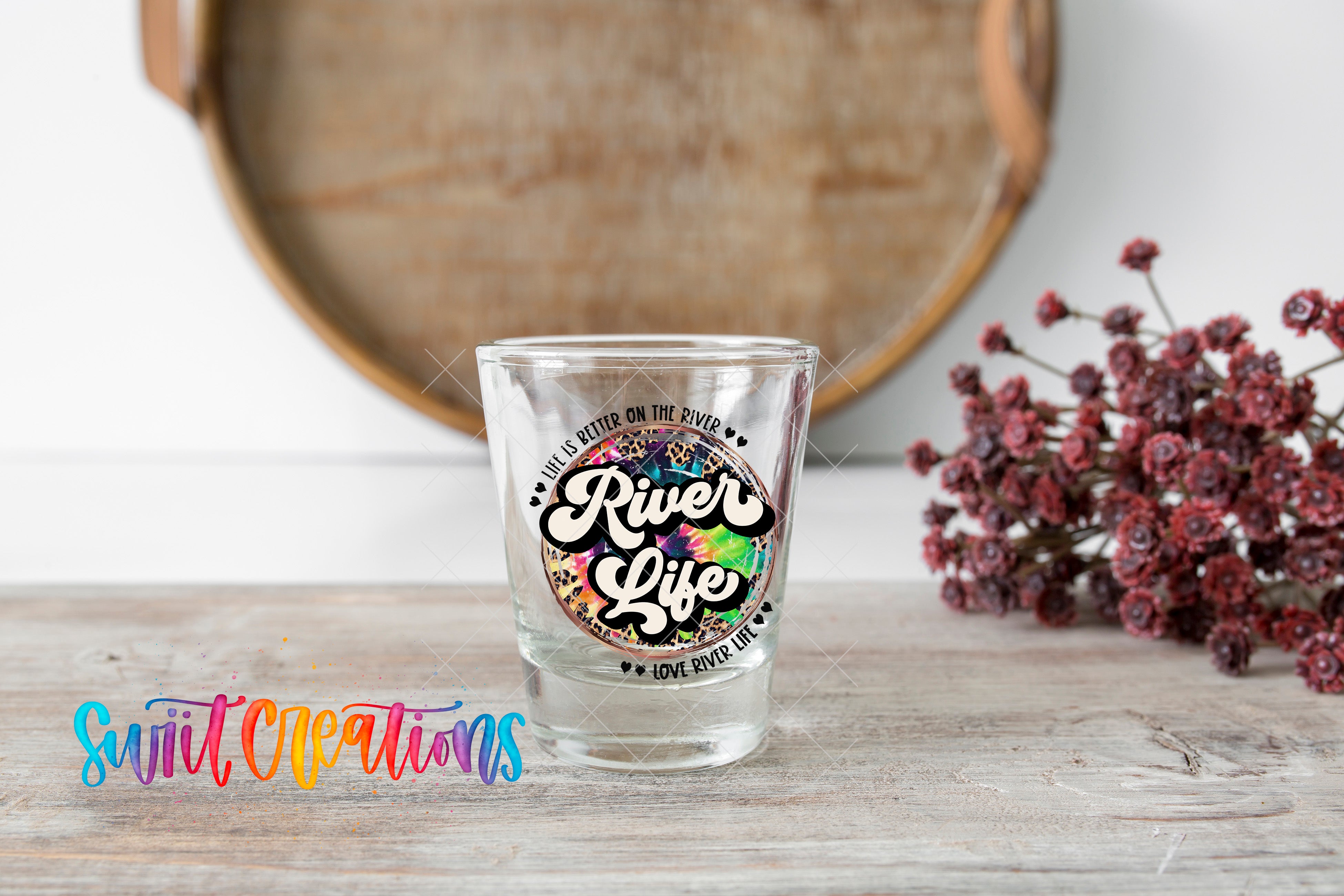 A shot glass with a colorful design and text on it, placed on a wooden surface next to a bunch of red flowers.