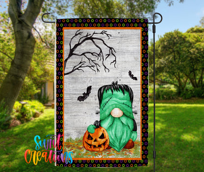 A Halloween-themed garden flag featuring a green gnome holding a carved pumpkin, with a spooky tree and bats in the background.