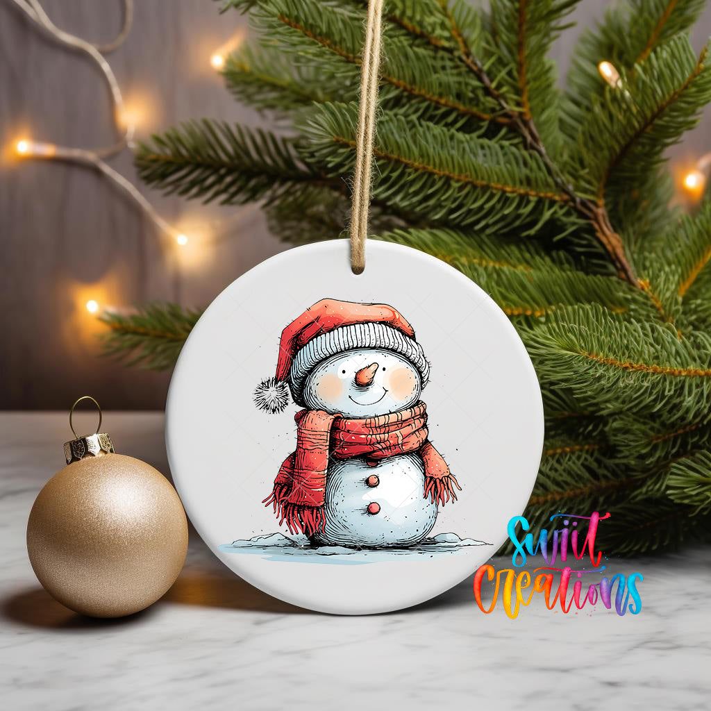 A white ceramic ornament featuring a snowman wearing a red scarf and hat, with a Christmas tree and twinkling lights in the background.