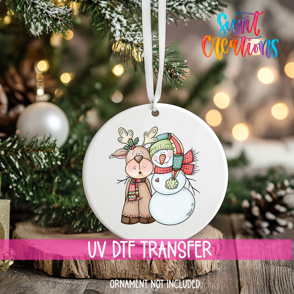 A white ornament with a deer and a snowman design hangs on a Christmas tree, with a pink ribbon around it.