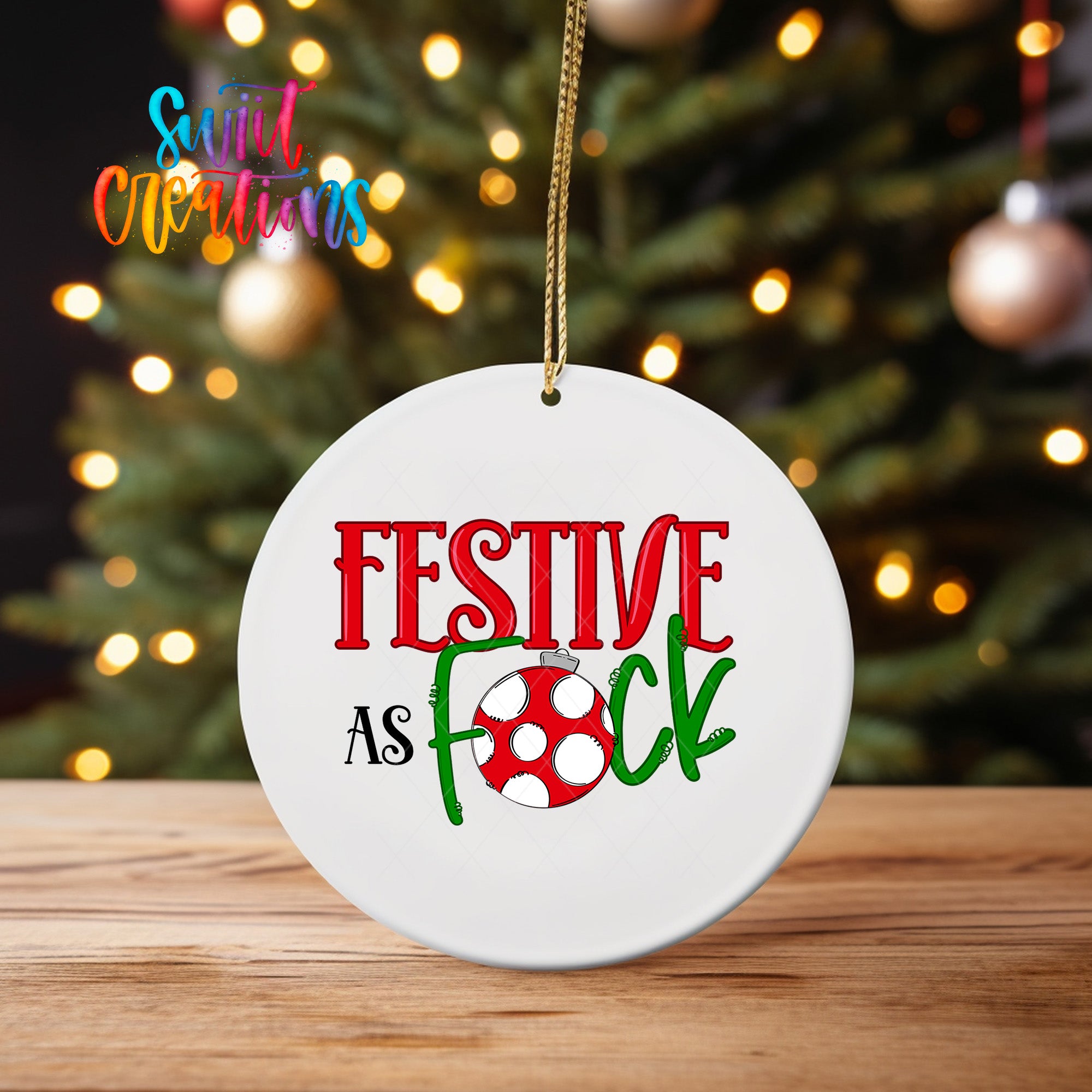 A white ornament with the text "FESTIVE AS FOCK" and a red and green design, hanging on a string in front of a blurred Christmas tree with colorful lights.