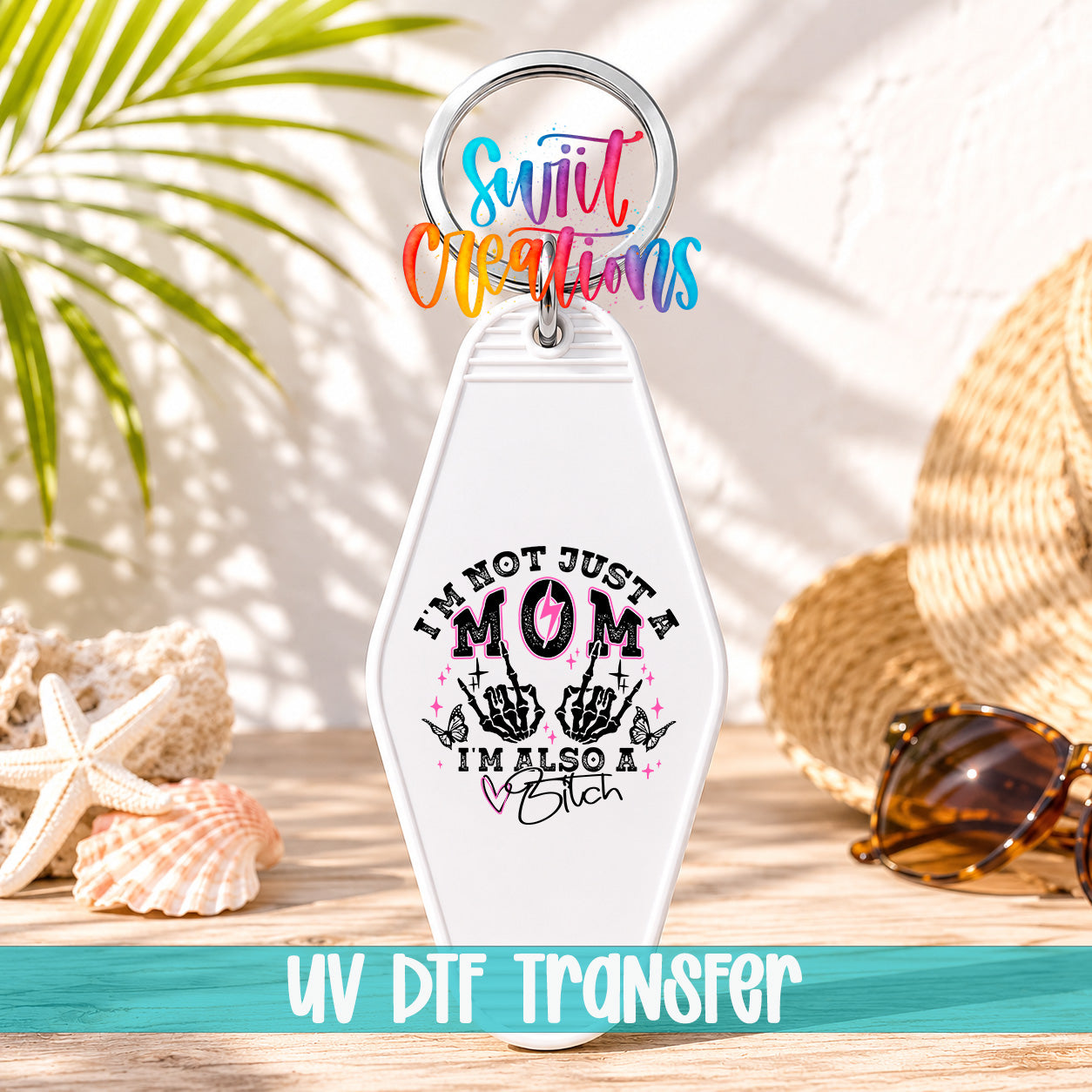 A keychain with a humorous text that reads "I'm not just a mom, I'm also a bitch" is displayed on a wooden surface, surrounded by sunglasses and a straw hat.