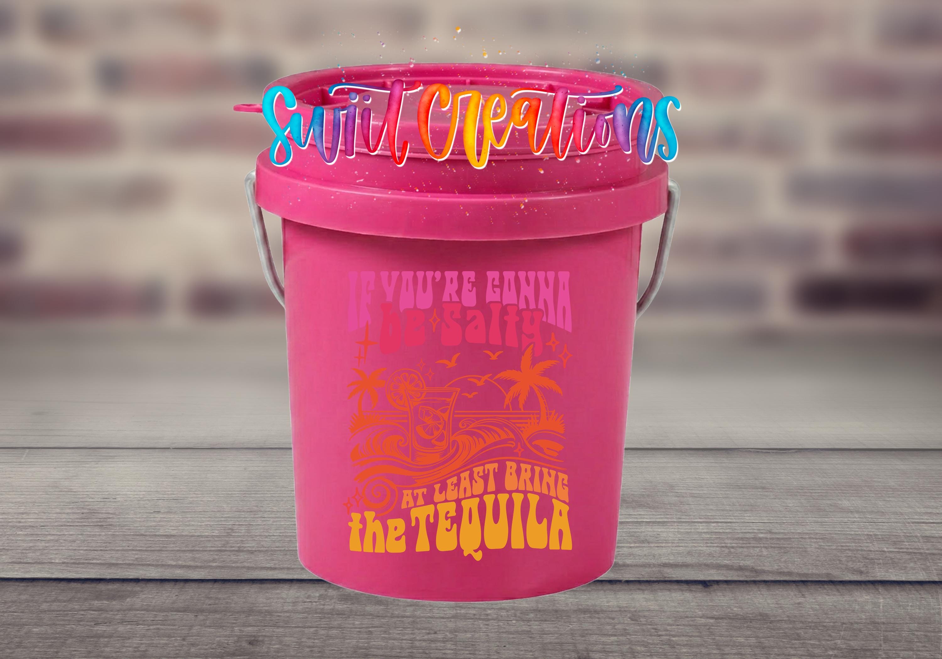 A pink bucket with a colorful, whimsical design and text on it, placed on a wooden surface against a blurred brick wall background.