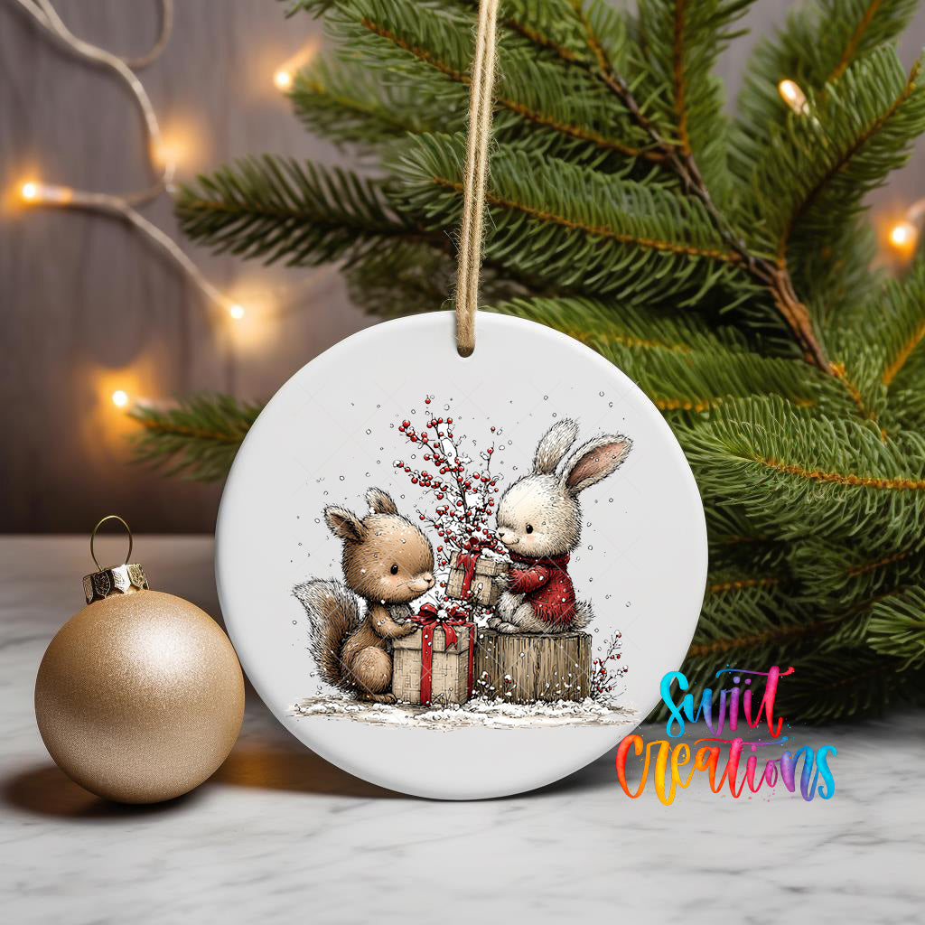 A white ceramic ornament featuring two cartoon rabbits, one wearing a red sweater, sitting on a wooden stump and holding a gift box, surrounded by snow and berries. The ornament is hanging from a string, and there is a gold ornament and a Christmas tree in the background.