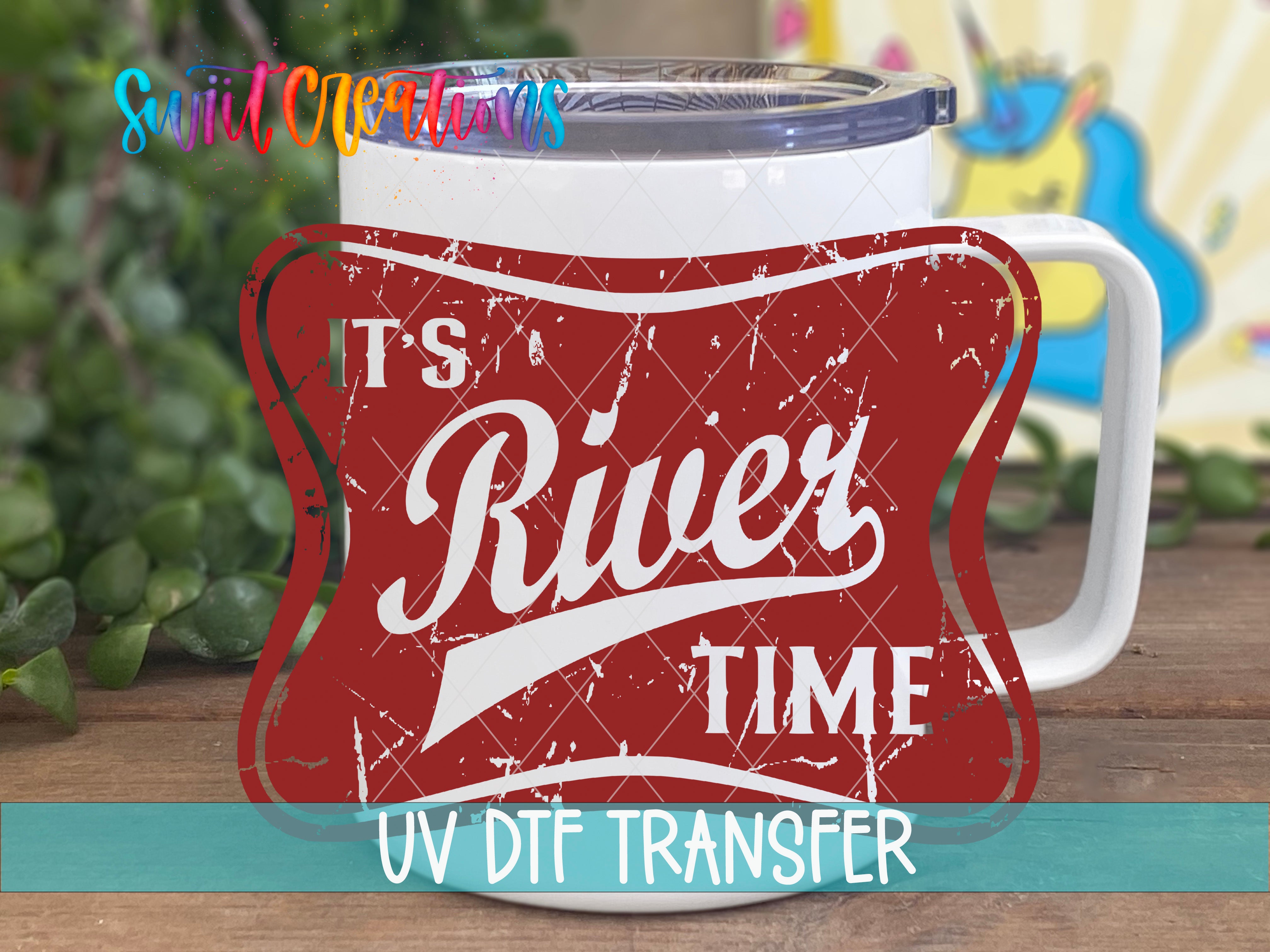 A white mug with a red and white label that says "River Time" and "UV Dtf Transfer" printed on it, placed on a wooden surface with green leaves in the background.