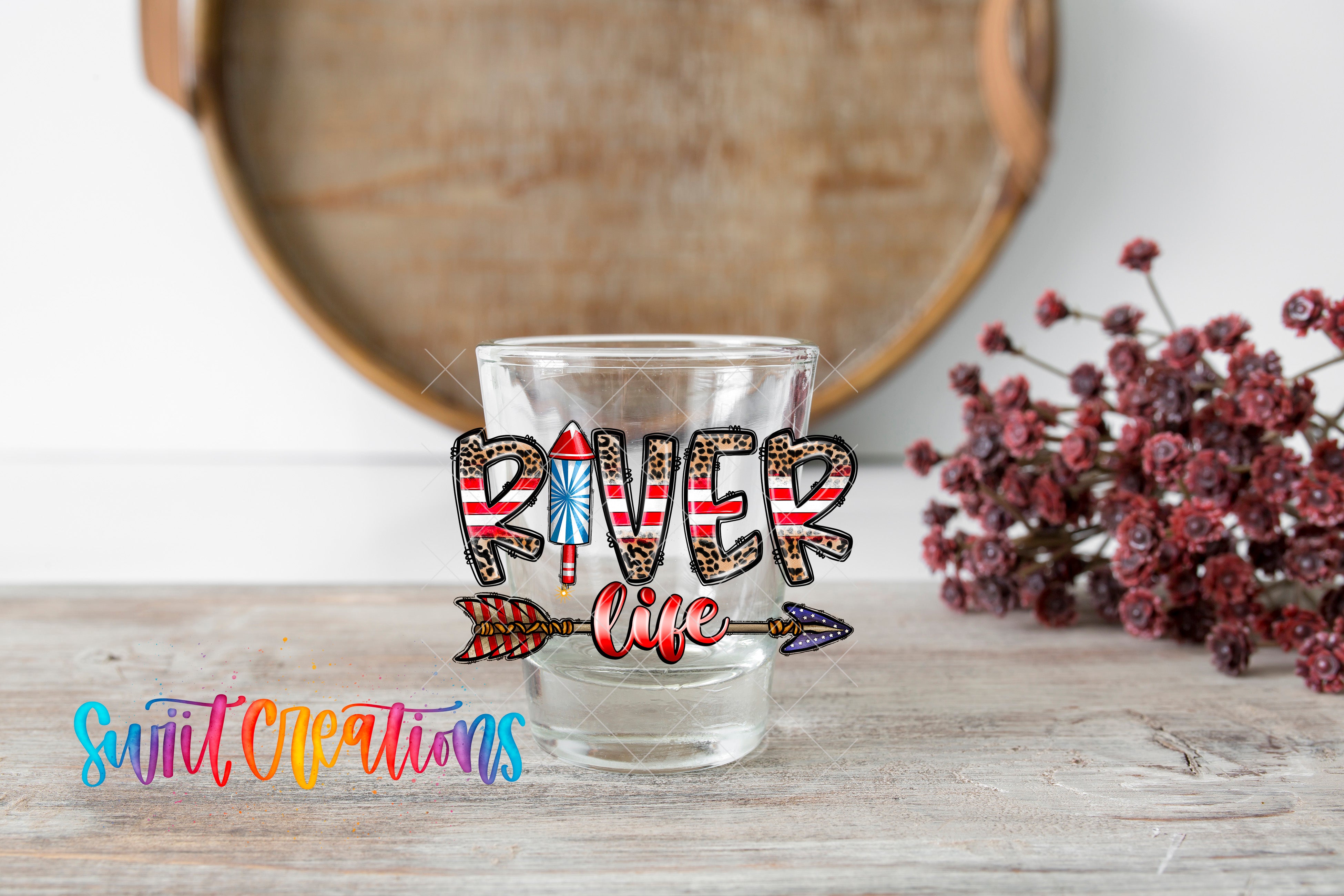 A glass with a decorative design featuring the word "RIVER" and an arrow, placed on a wooden surface with a floral arrangement in the background.