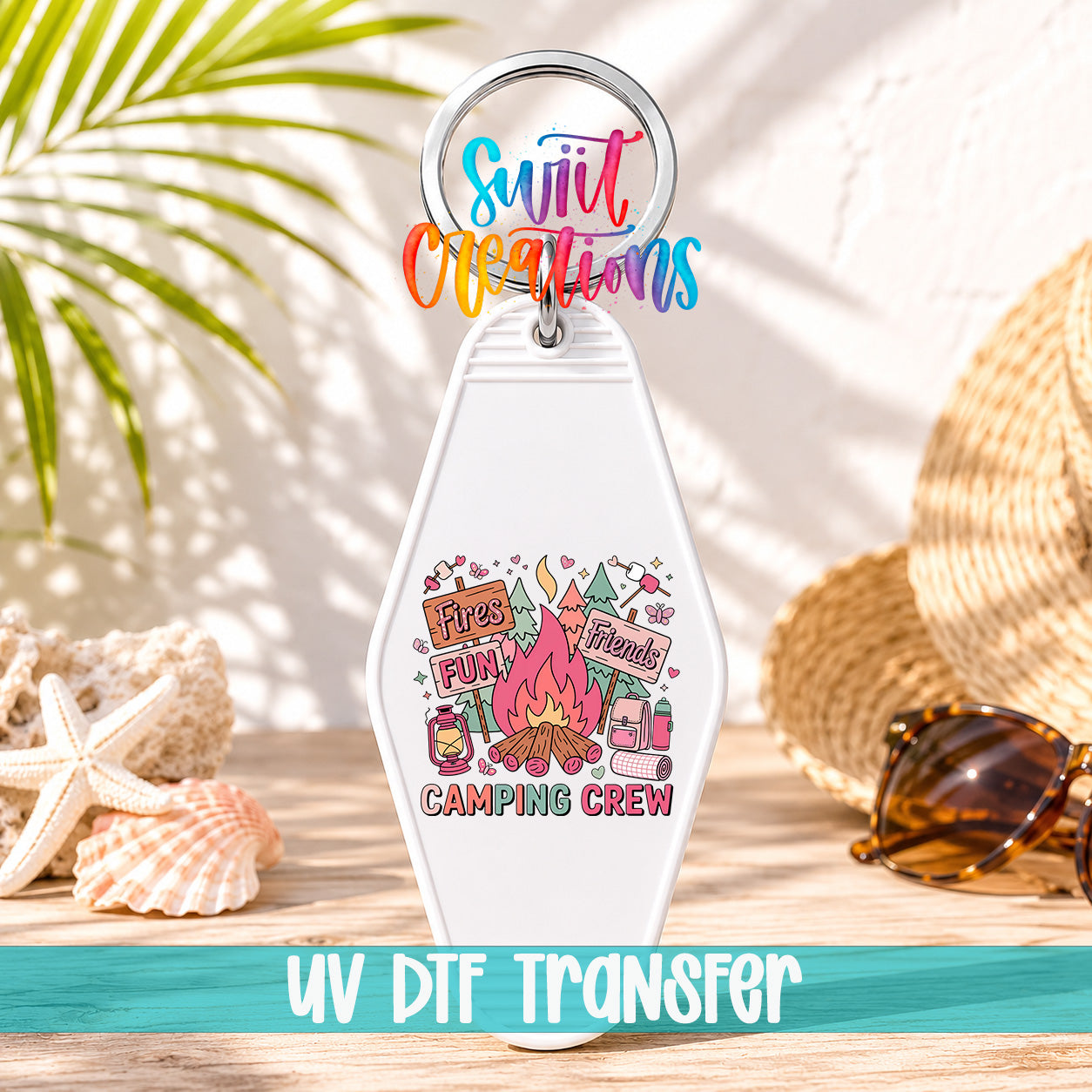 A keychain with a colorful design featuring camping-related items, such as a campfire, tent, and camping equipment, along with the text "Camping Crew" and "Fires Fun Friends". The keychain is placed on a wooden surface, with sunglasses and a straw hat visible in the background.