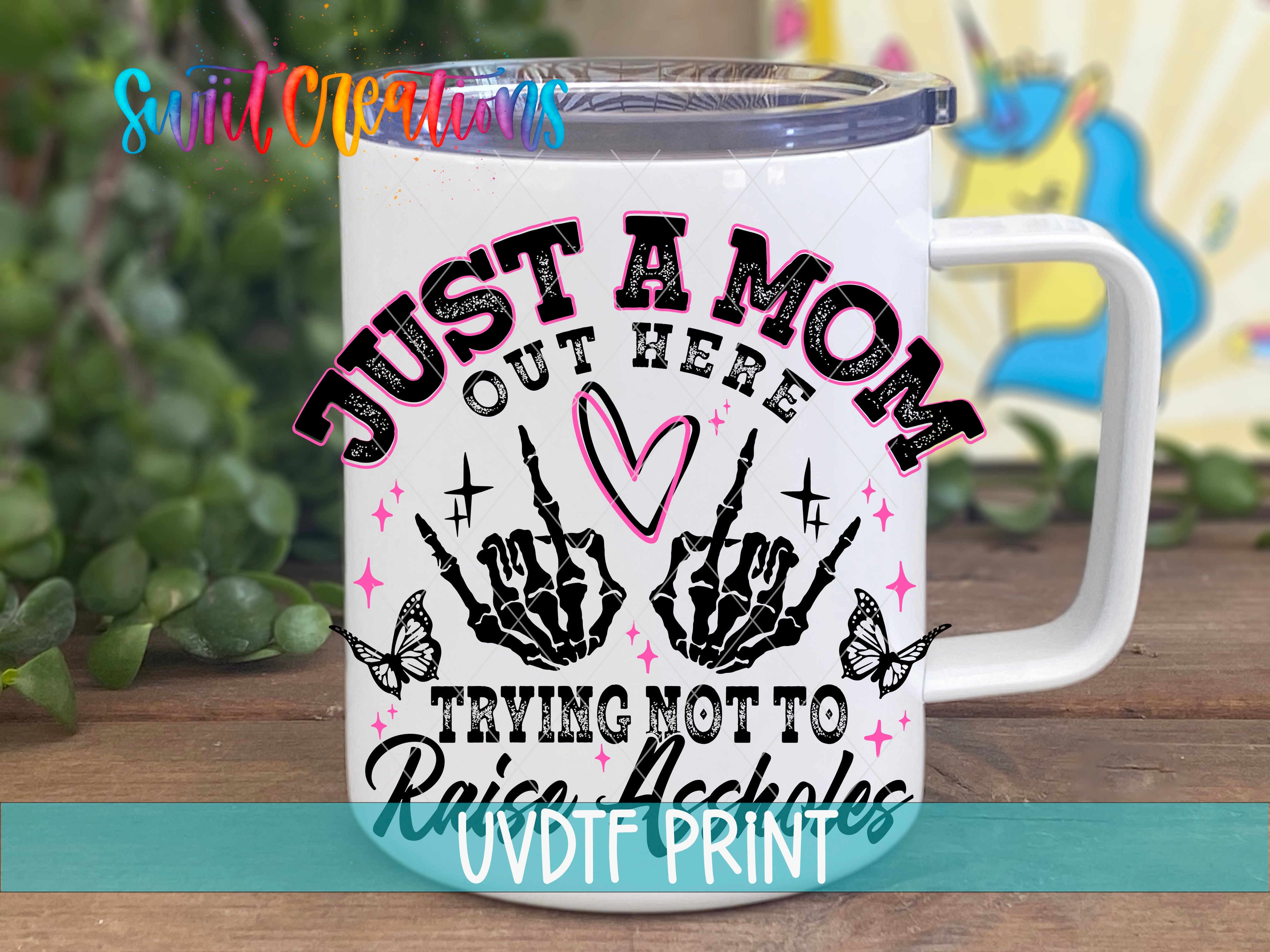 A white mug with a colorful design featuring a skeleton hand and the text "Just a mom out here trying not to raise hell" printed on it.