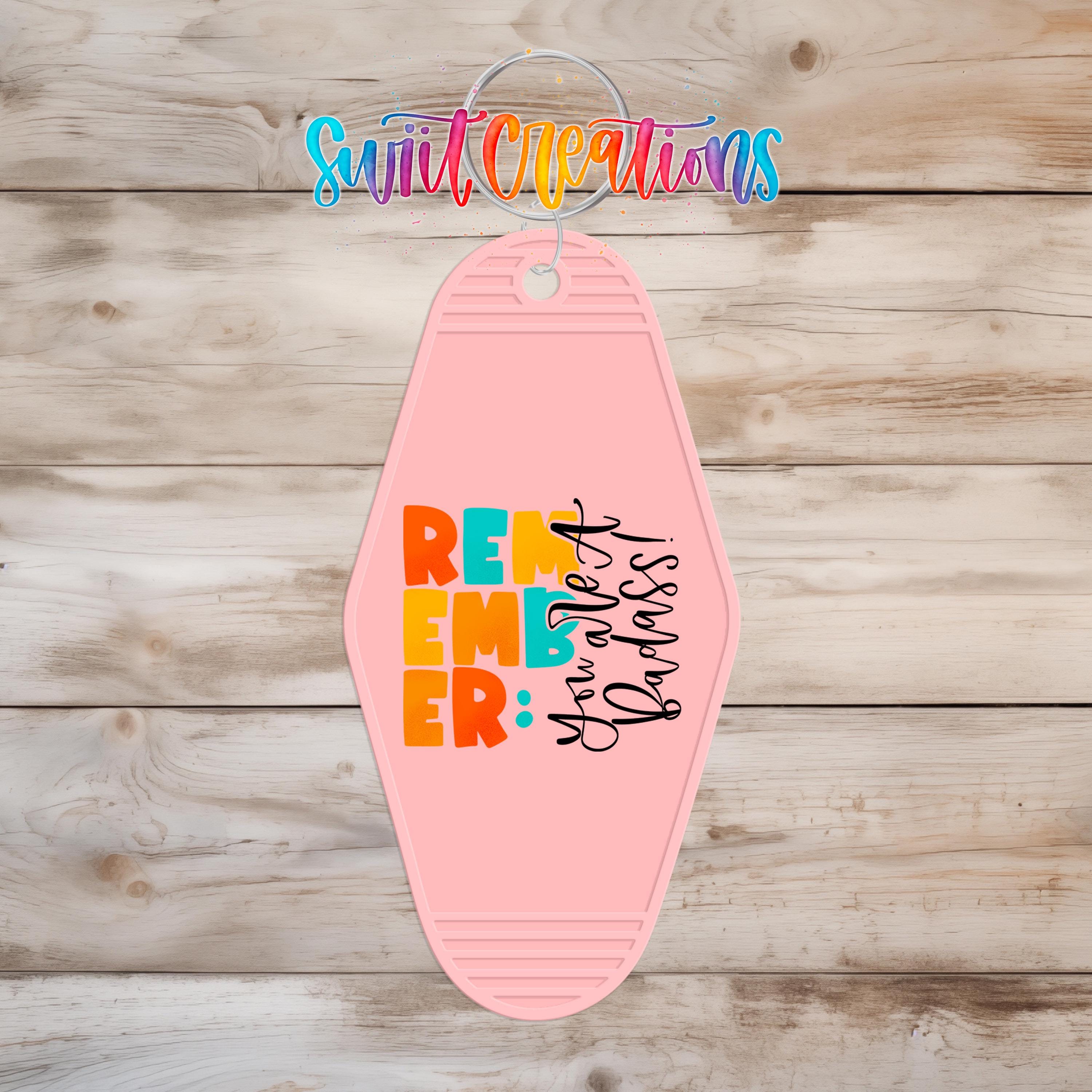 A pink keychain with the text "Remember Badass" printed on it, hanging on a wooden surface.