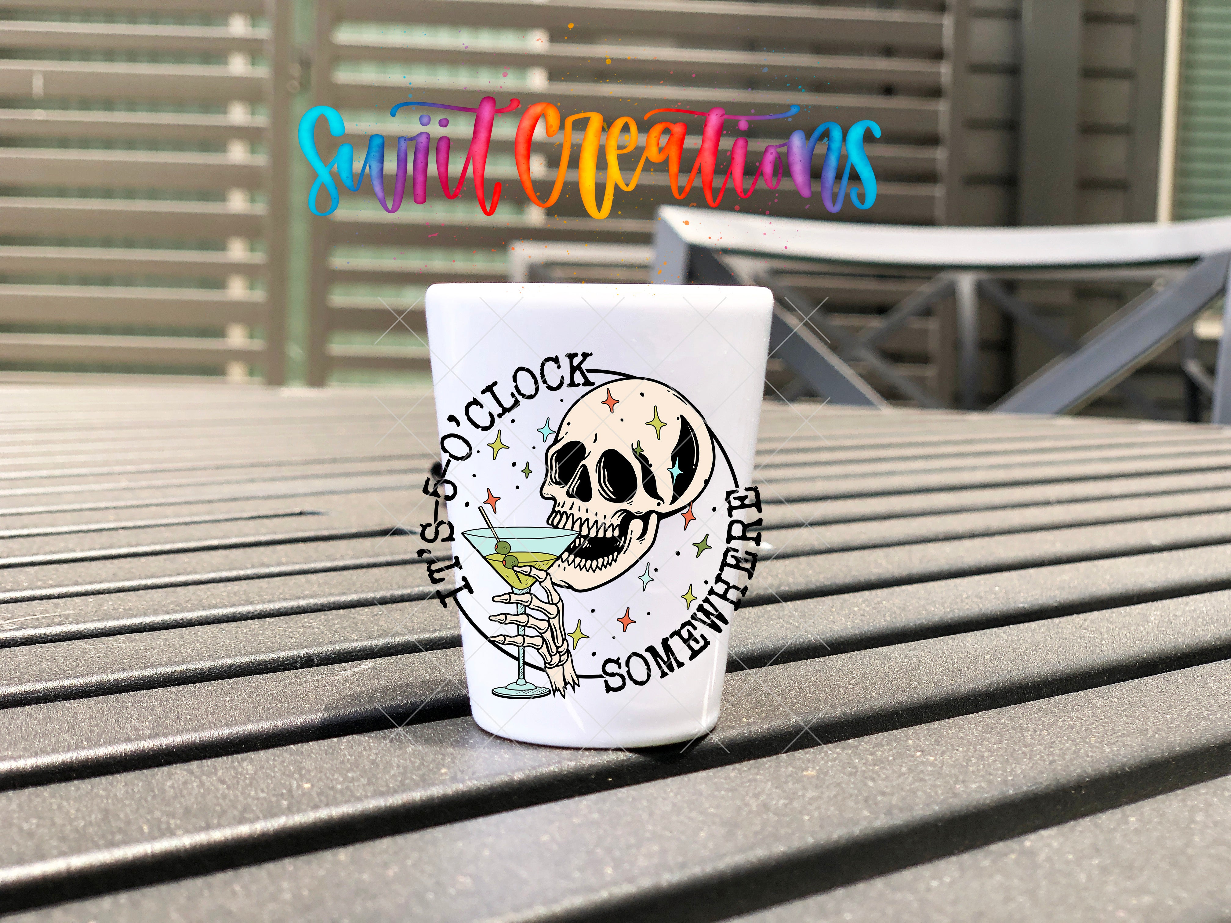 A white shot glass with a skull design and the text "It's 5 o'clock somewhere" printed on it, placed on a wooden surface.