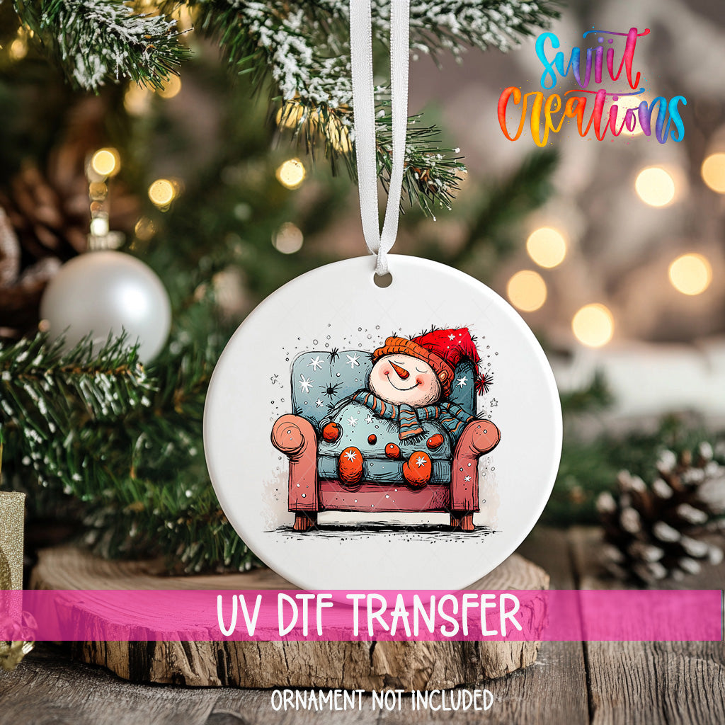 A white ornament with a snowman sitting in a chair is hanging on a Christmas tree.